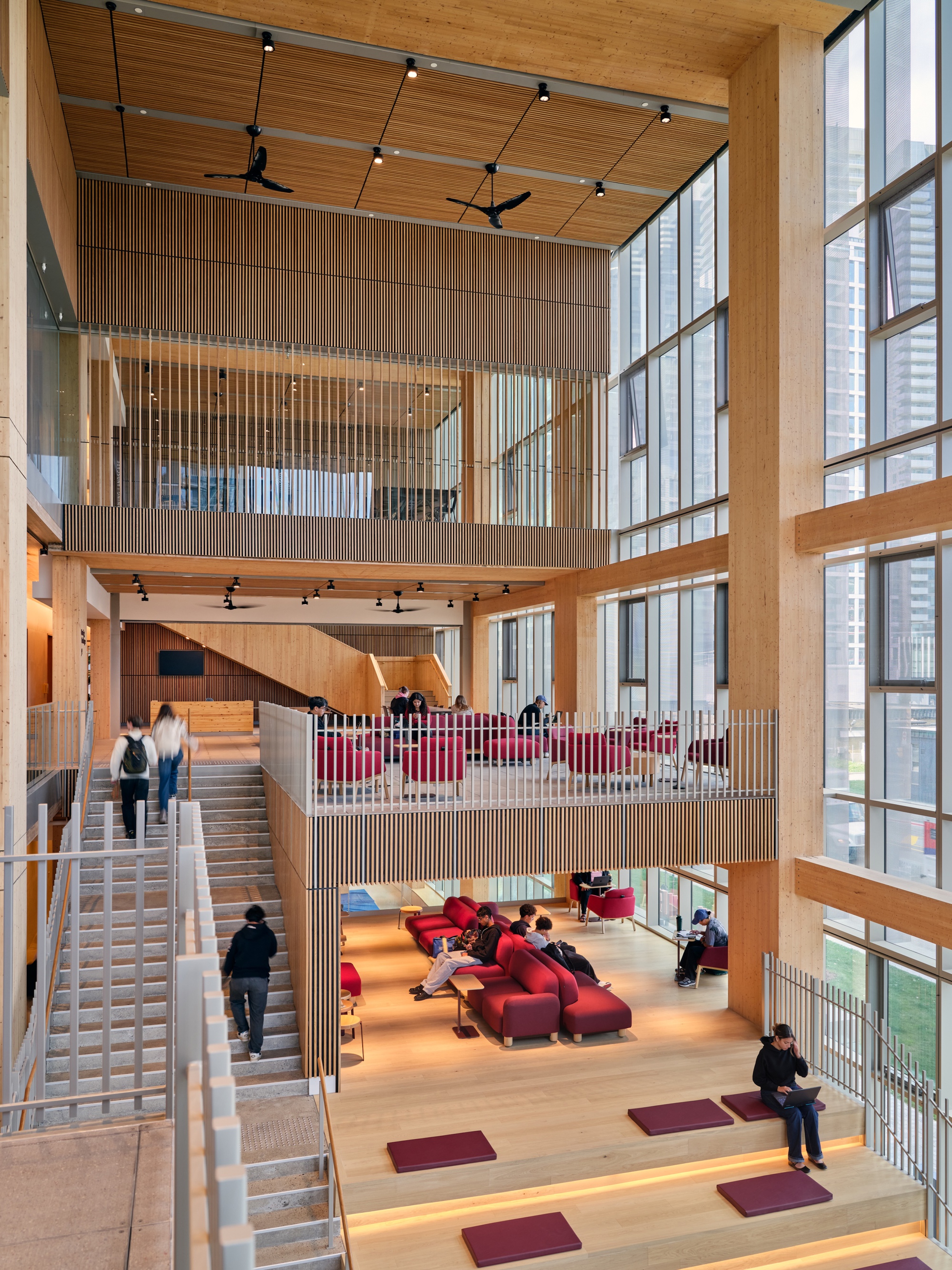 Triple-height atrium with timber screens, stepped platforms, and people gathering on red carpet squares
