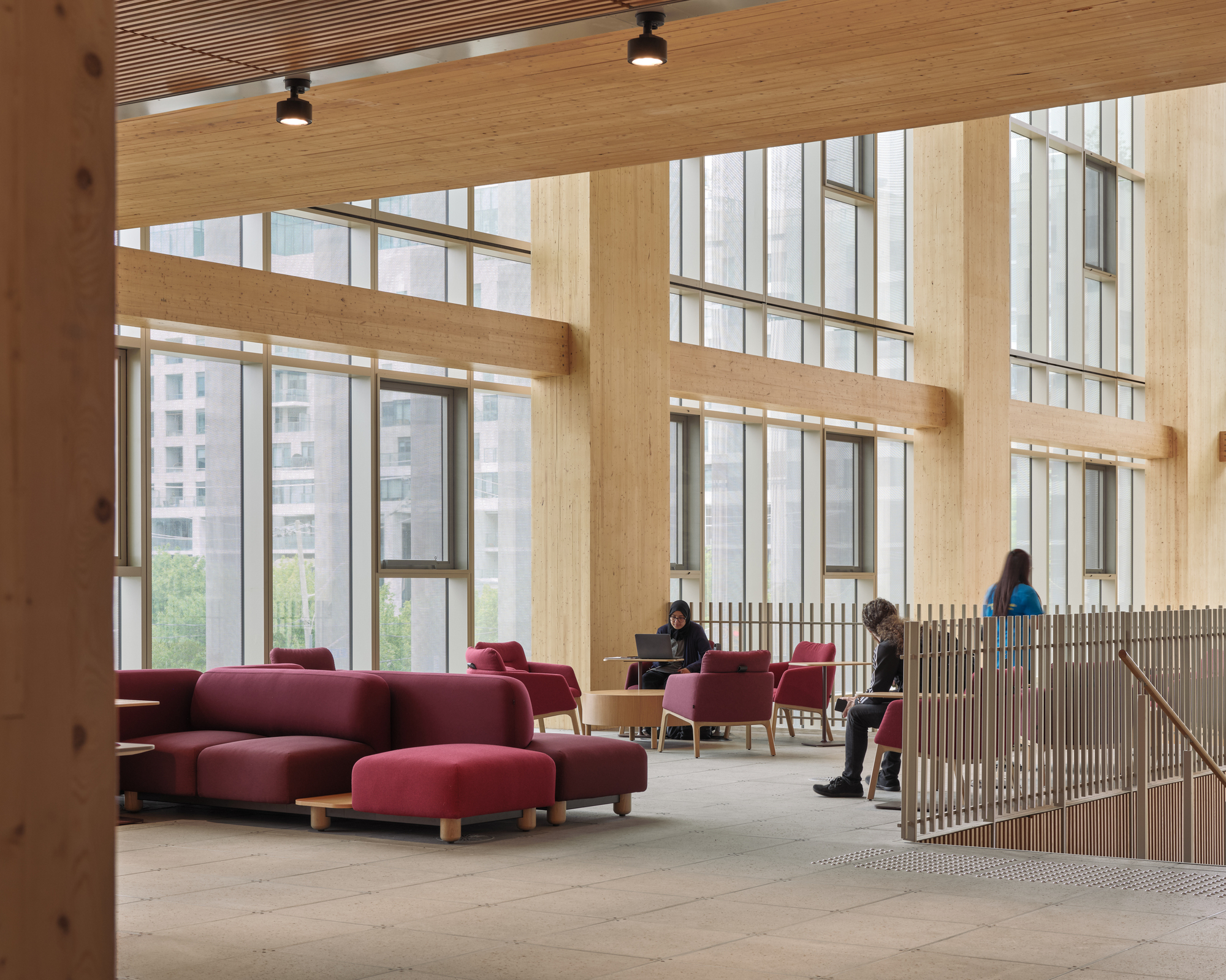 Upper-level lounge with burgundy modular seating, wood slat railing, and tall windows framing buildings beyond