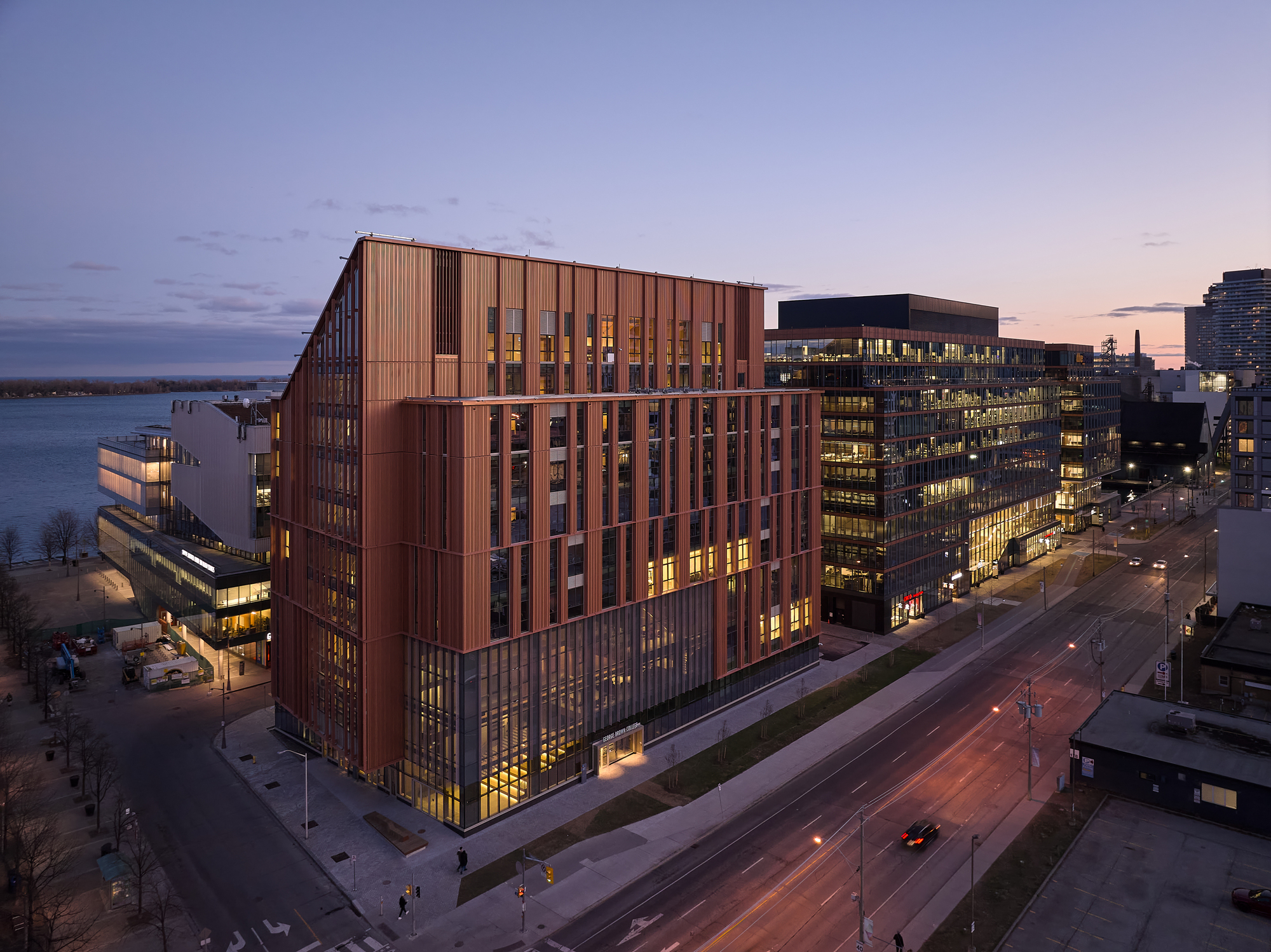 Aerial view of the copper-clad facade with vertical fins illuminated at dusk beside a waterfront boulevard