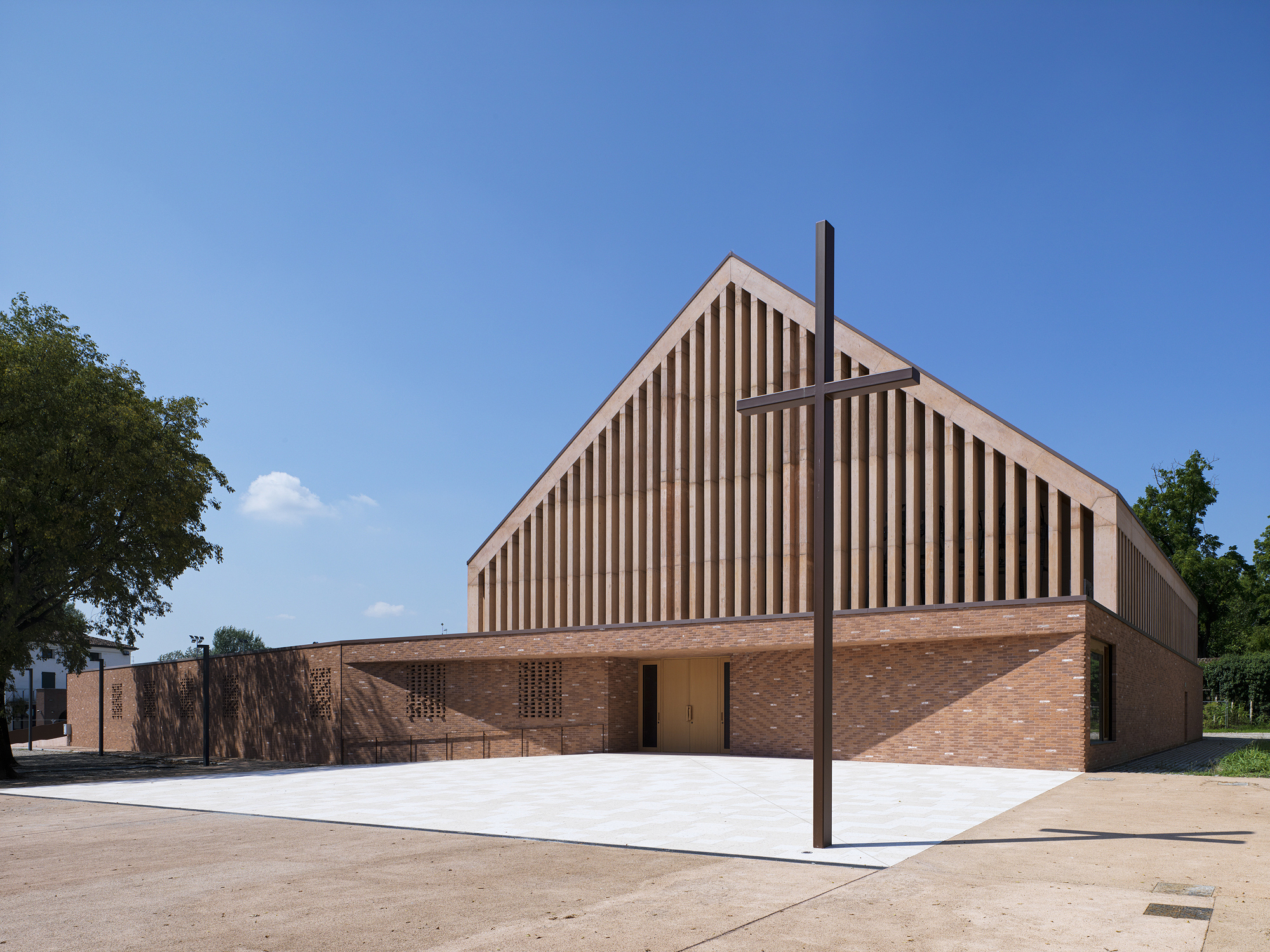 Gabled volume with vertical timber louvers above a brick base and a steel cross in the forecourt
