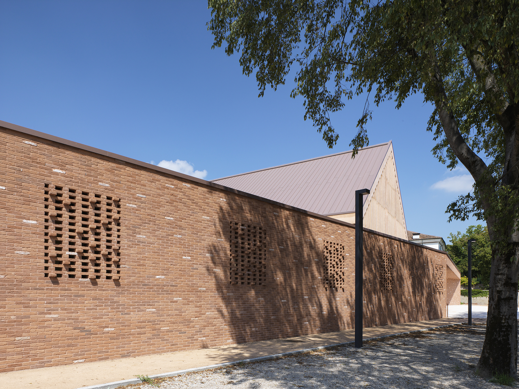 Perforated brick facade with angular gable roof under a clear blue sky and tree shadow