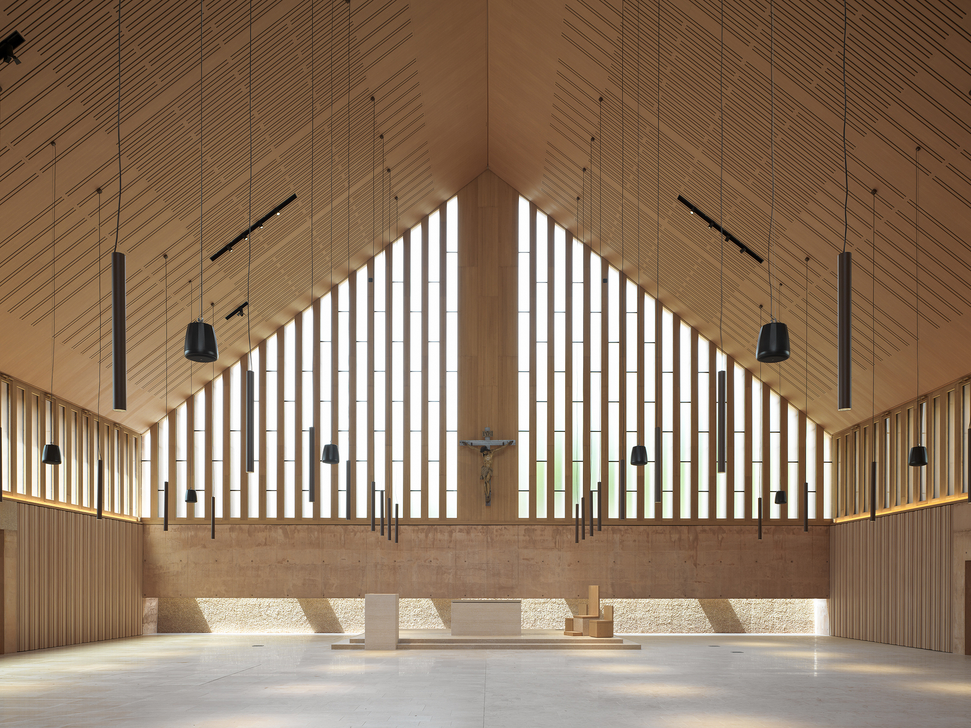 Altar wall with vertical timber screens, crucifix and suspended lighting beneath a pitched wood ceiling