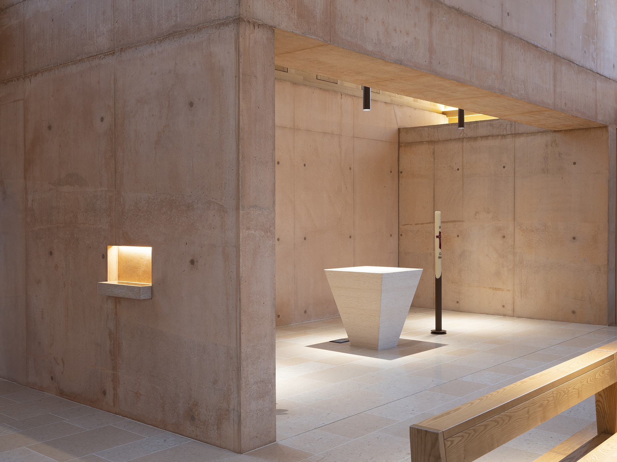 White stone baptismal font on a raised platform beneath exposed concrete beams and recessed wall niches