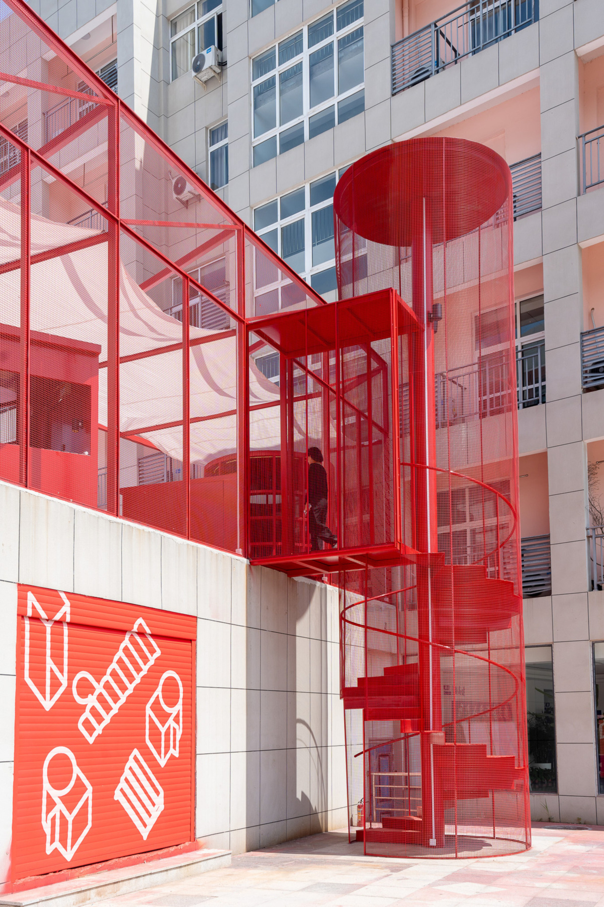 Exterior red spiral staircase enclosed in perforated metal mesh climbing the tiled facade