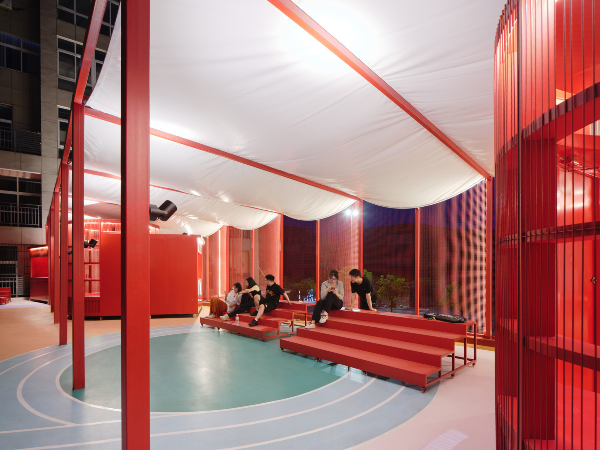 Covered pavilion with red steel columns, white fabric canopy and people seated on stepped benches at dusk