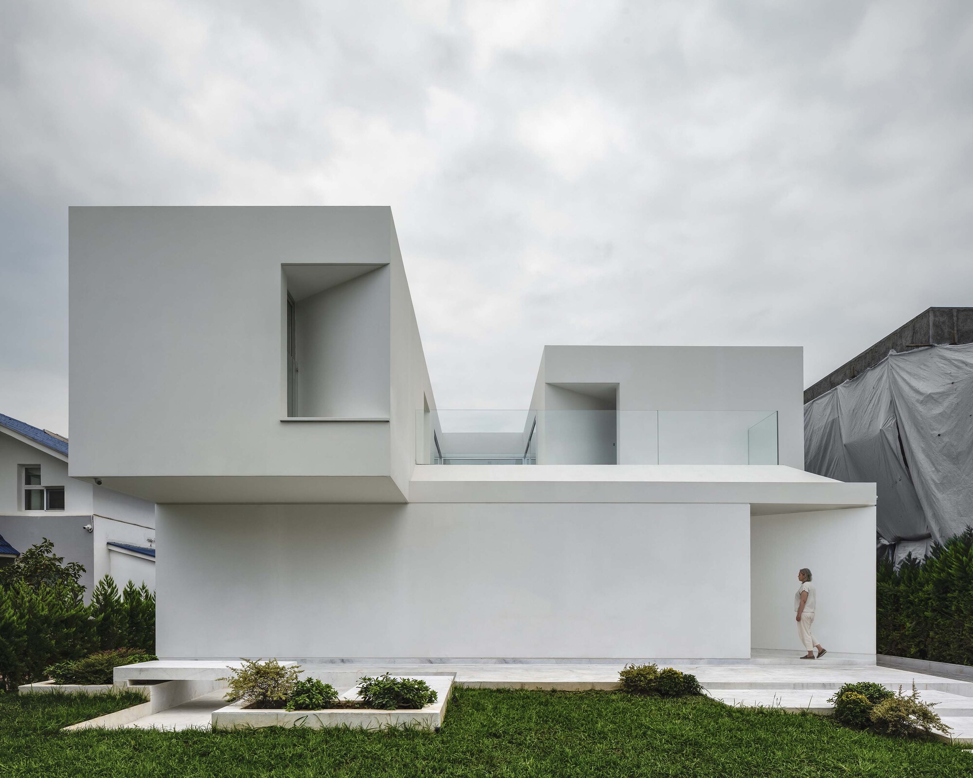 White rendered facade with cantilevered volumes and recessed entrance where a figure stands under overcast skies
