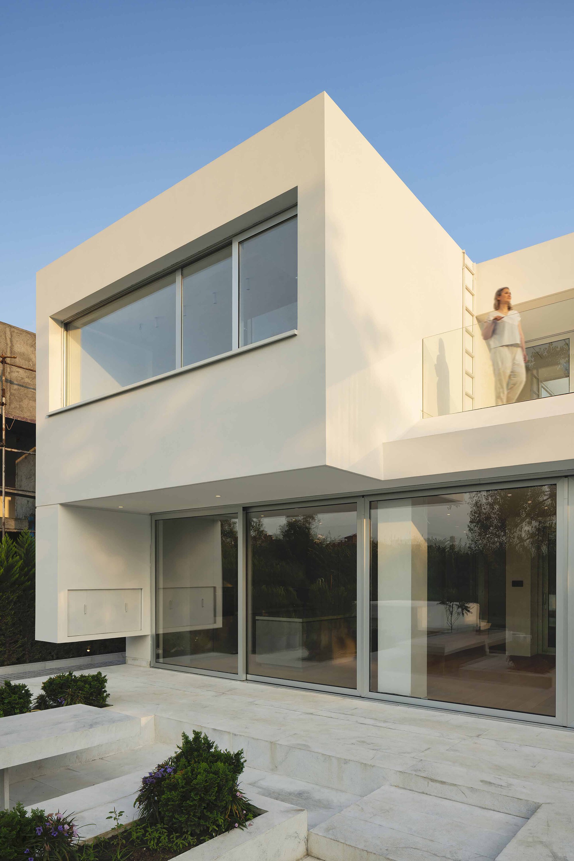 White rendered facade with full-height glazing and upper-level balcony at dusk with a figure visible