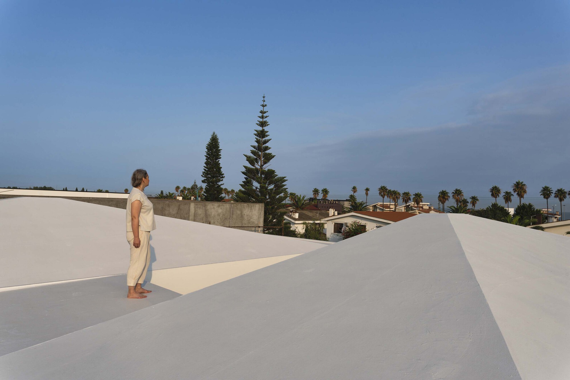 Rooftop terrace with white rendered volumes and a figure overlooking palm trees in the distance