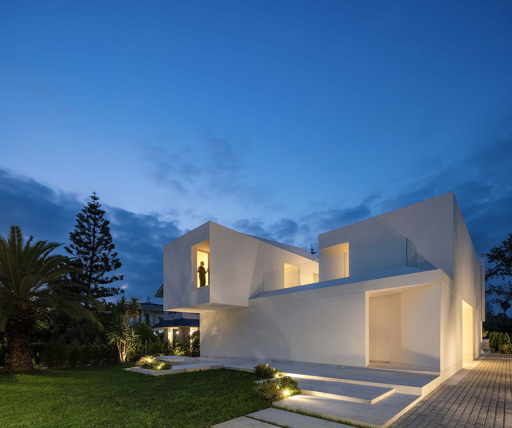White cubic volumes with illuminated openings and recessed glass at twilight beside a lawn