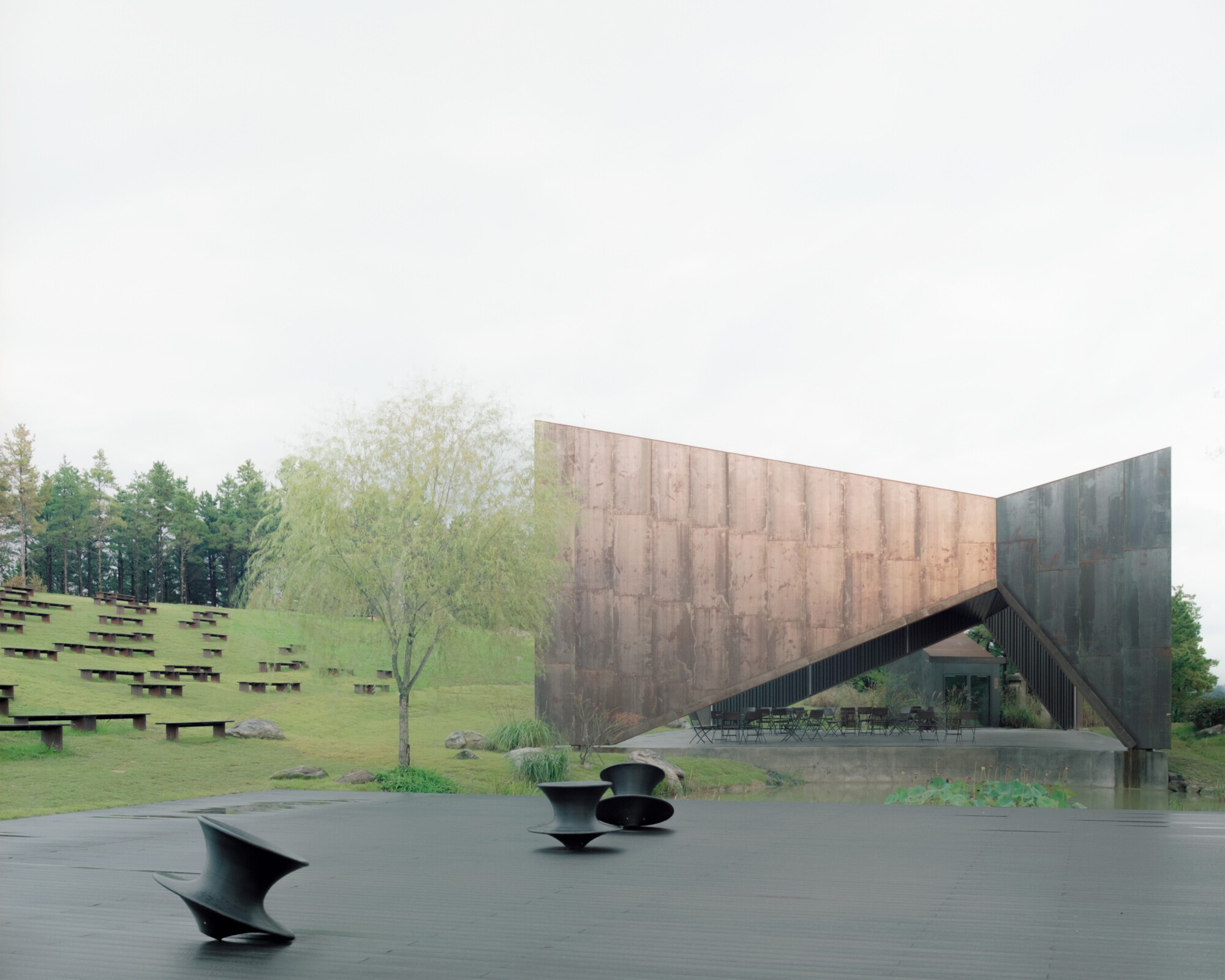 Weathered steel facade with sculptural metal benches on a wet plaza in spring