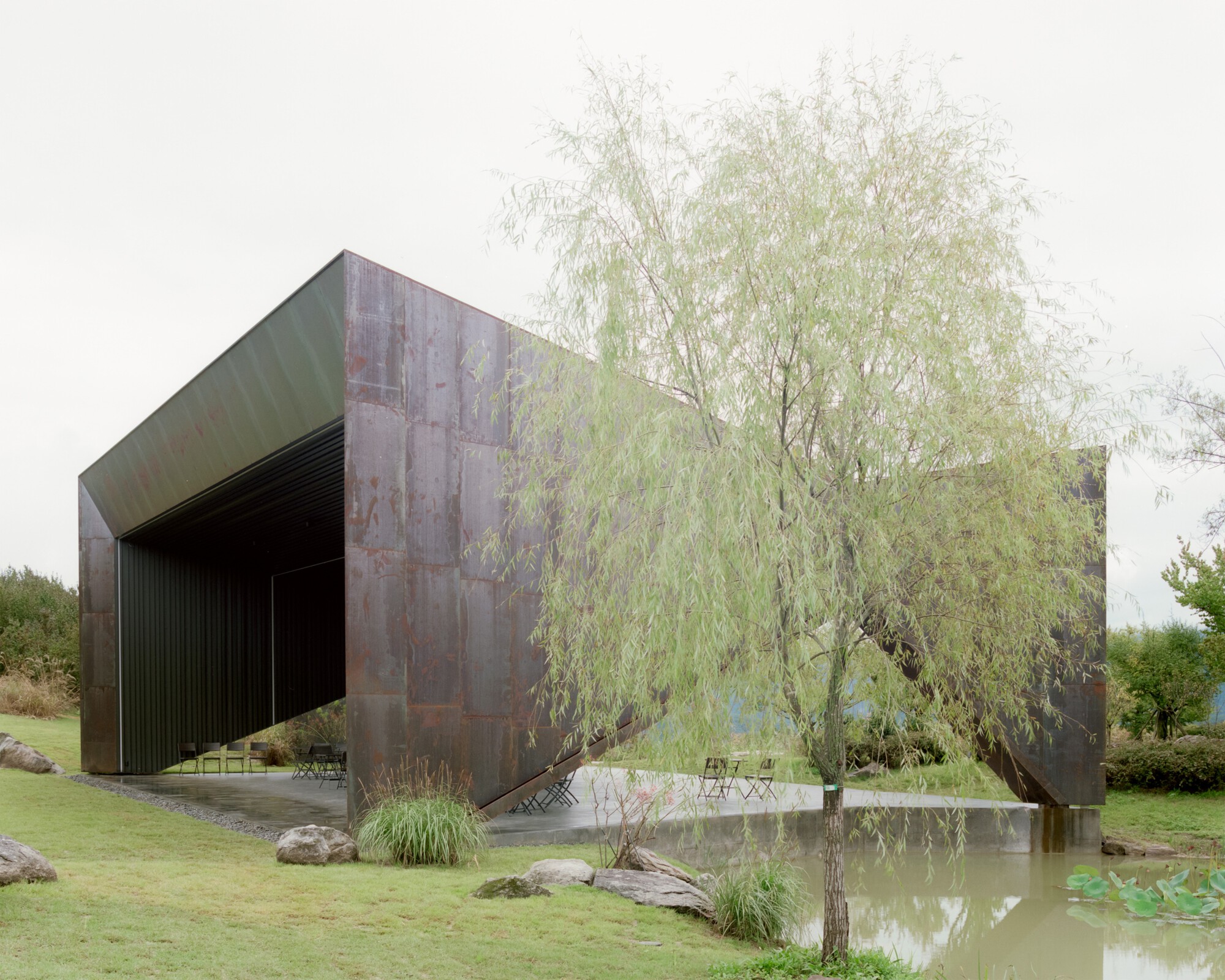 Corten steel exterior walls beside a shallow reflecting pool with willow tree and natural grasses