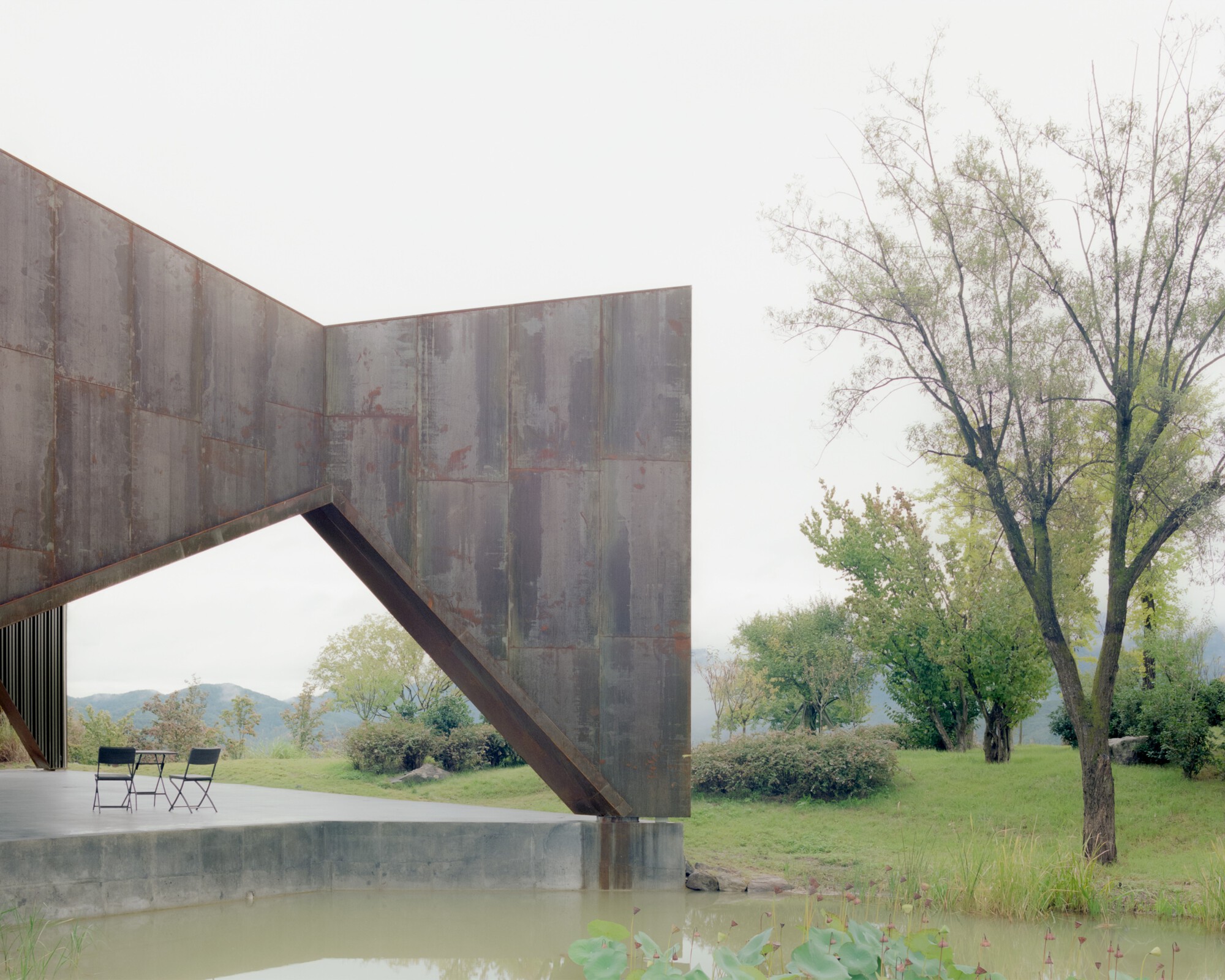 Angled steel support meeting the weathered panel facade above a concrete terrace and shallow pool