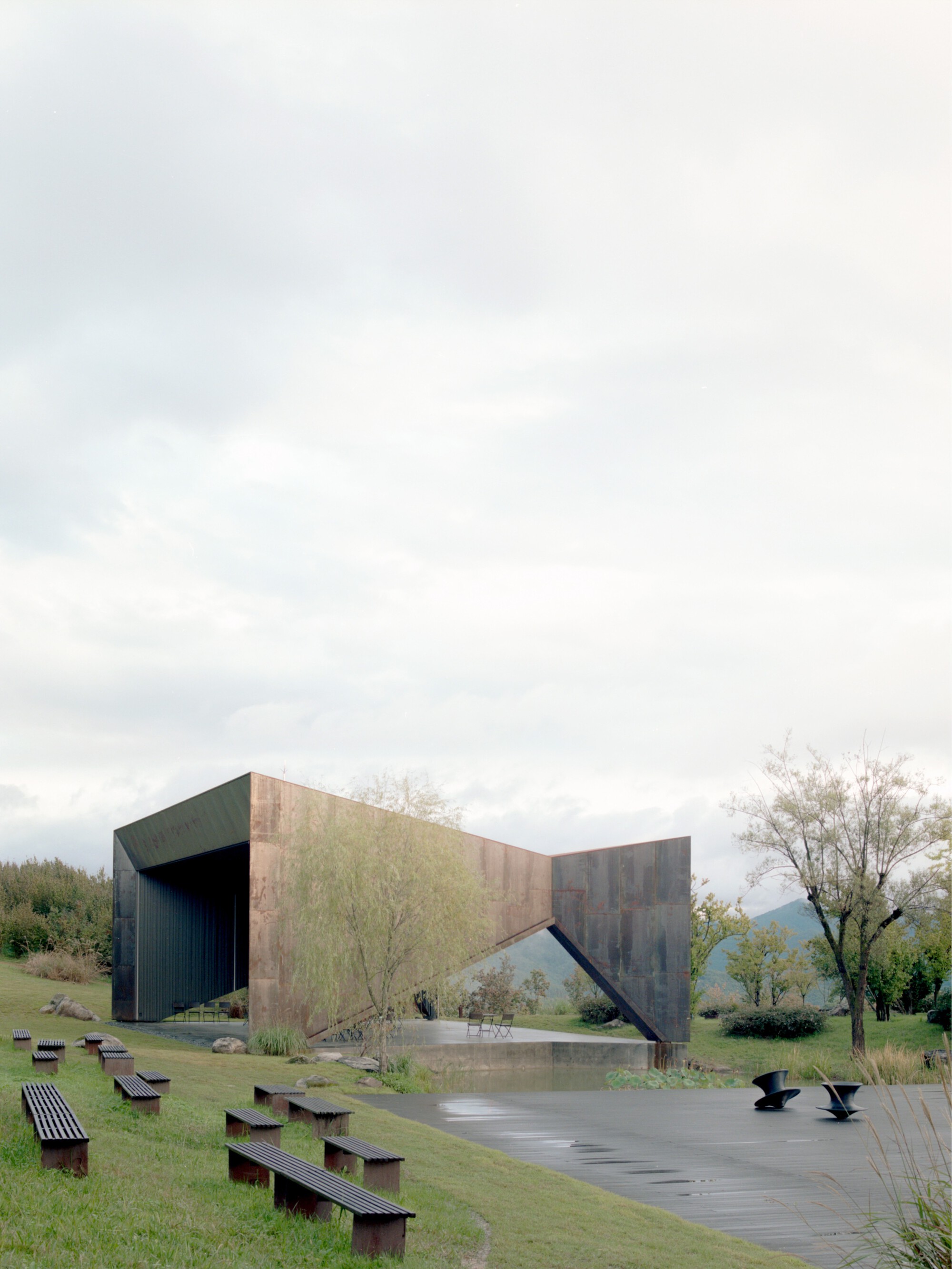 Side view of the folded steel structure with reflecting pool and benches under an overcast sky