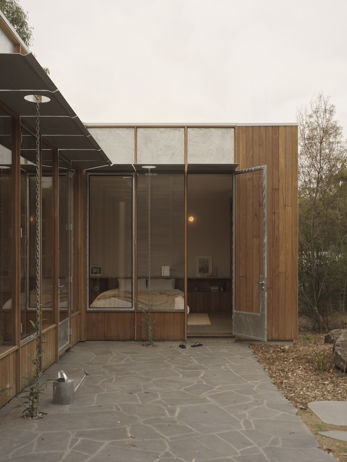 Covered exterior terrace with hanging swing seat and stone paving looking into a bedroom volume at dusk