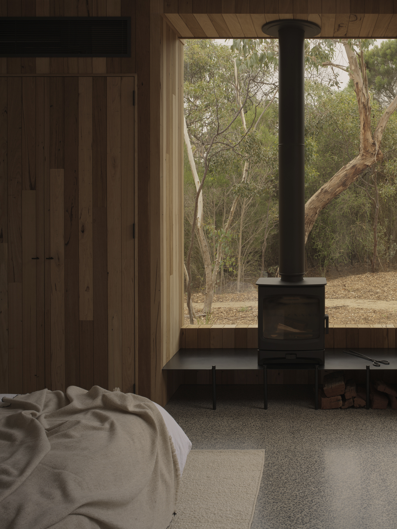 Interior view of freestanding black wood stove on steel plinth with timber wall and window overlooking trees