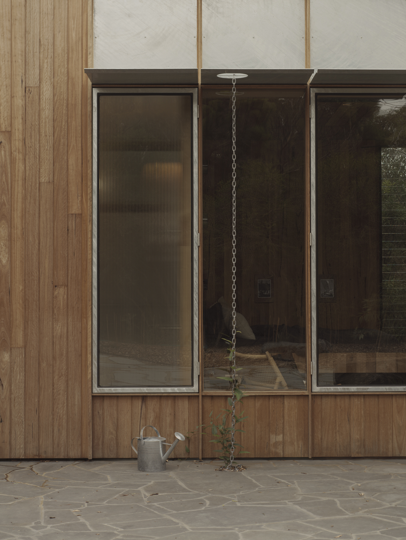 Timber-clad facade with tall glazed panels above a stone terrace with hanging plant and watering can