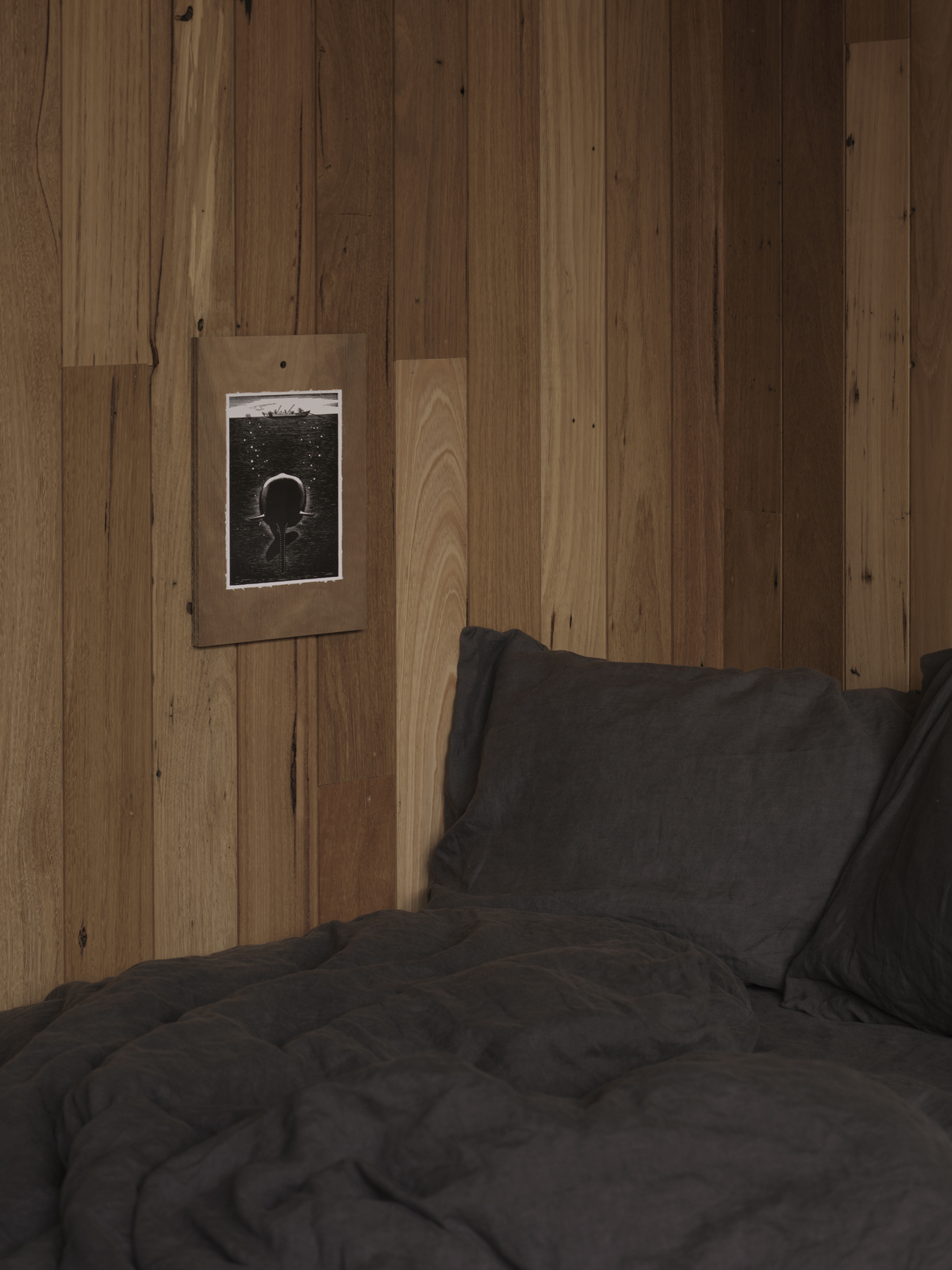Framed photograph mounted on vertical timber wall above dark grey bed linens in dim light