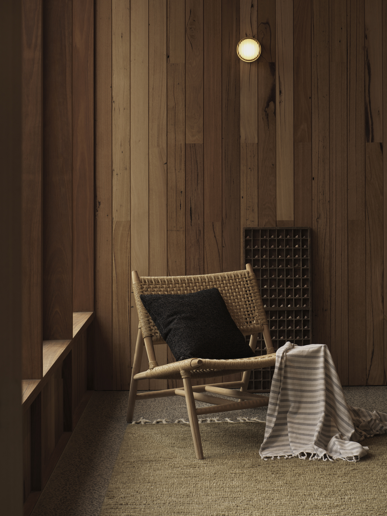 Woven lounge chair with black cushion against vertical timber-clad wall with a single ceiling light