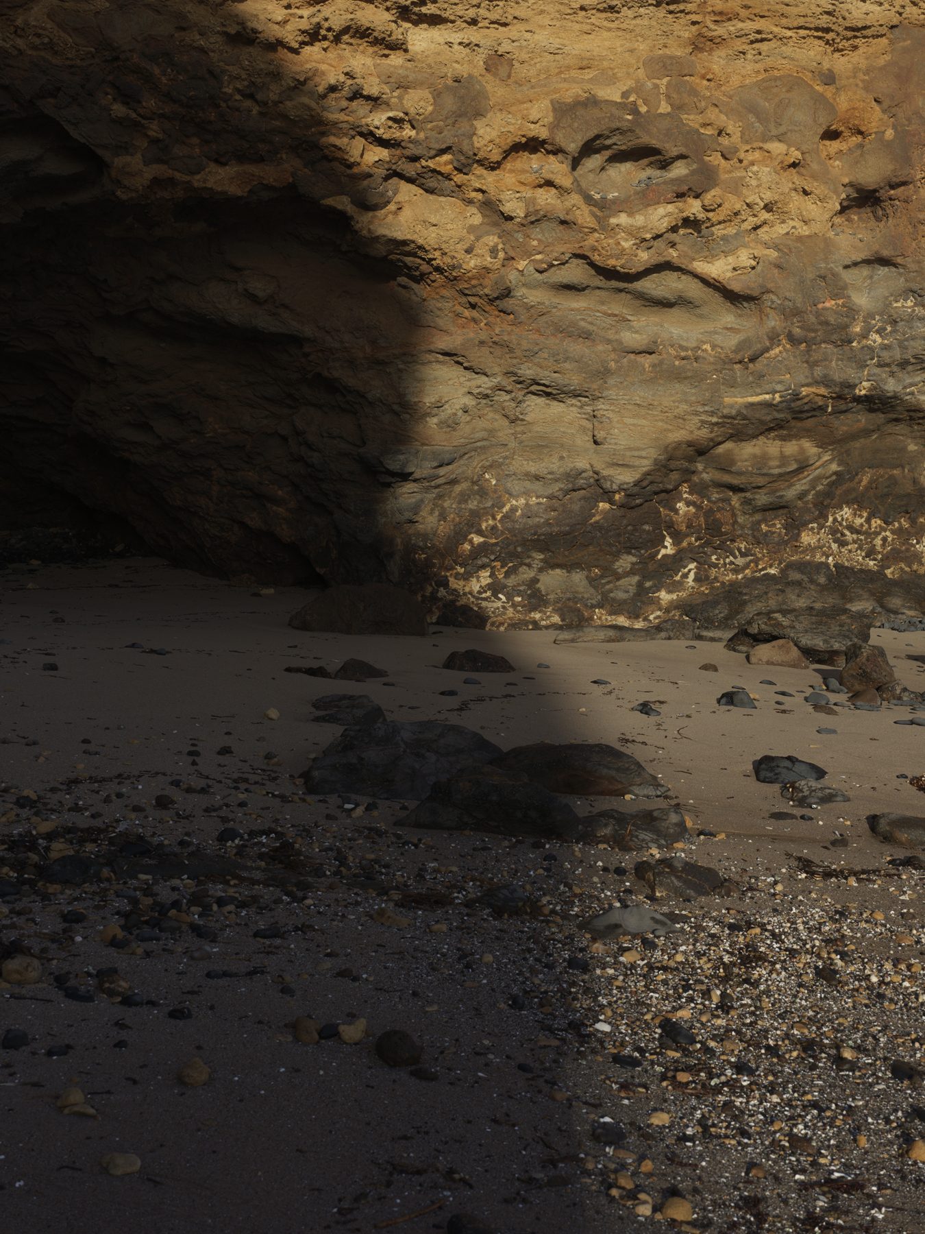 Natural sandstone overhang with layered sediment formations above a pebbled floor in shadow