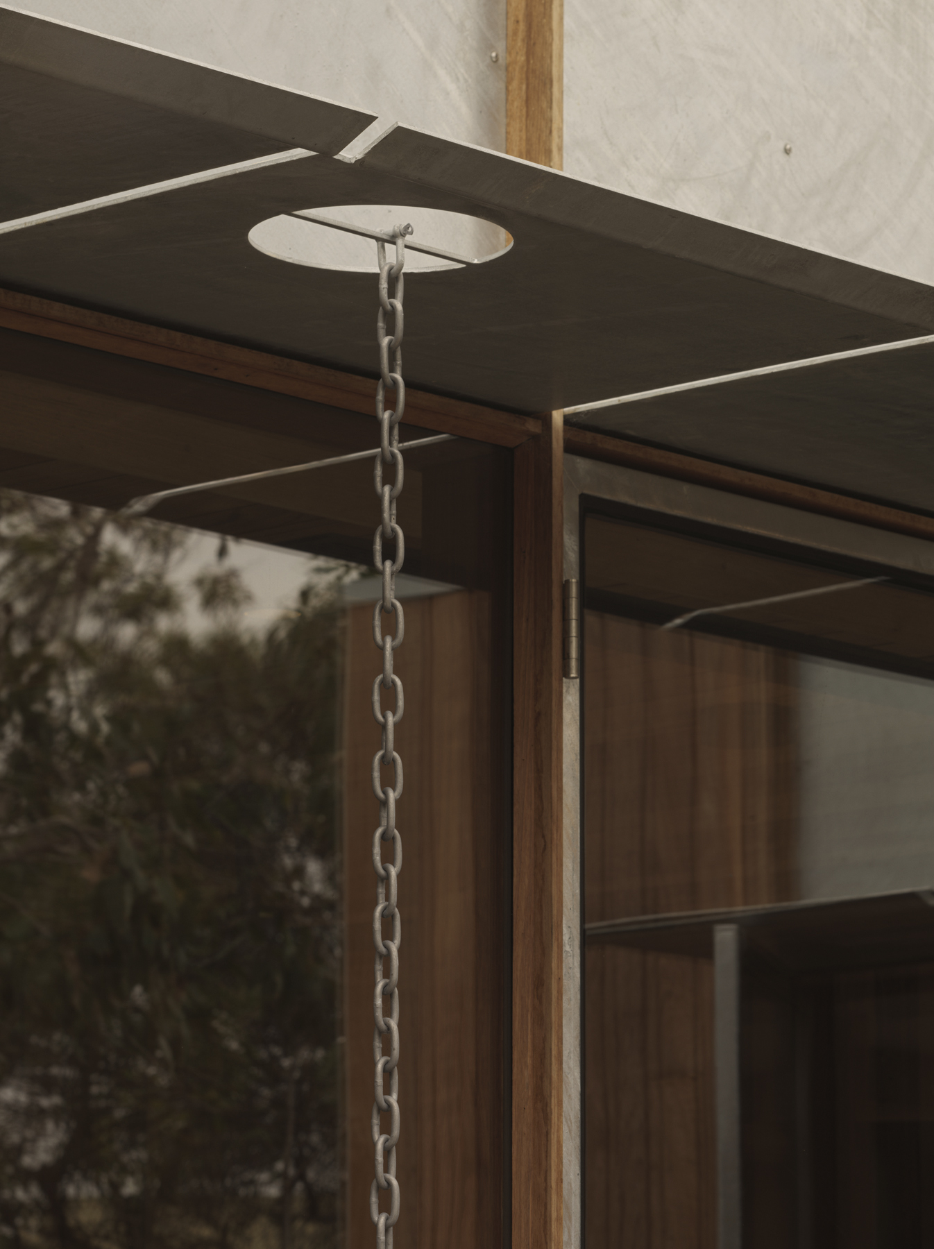 Rain chain suspended from timber-framed eave with concrete soffit overlooking vegetation