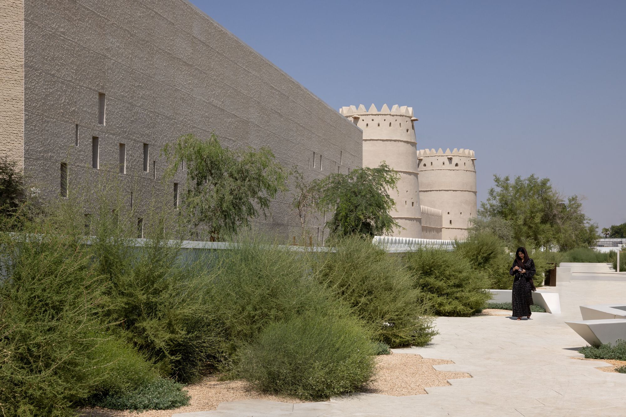 Perforated stone facade beside a gravel path with planted shrubs and a person walking in the distance