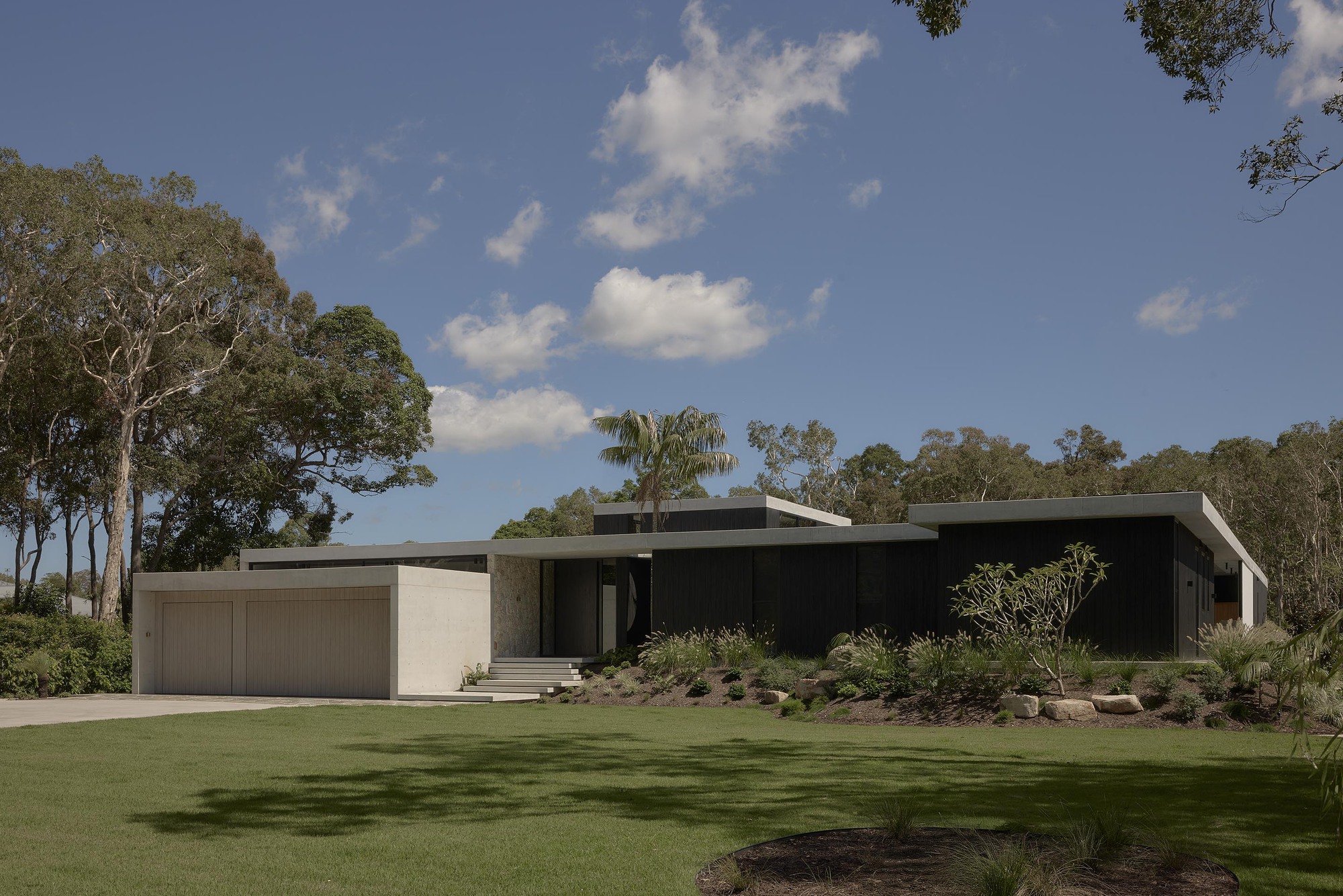 Long horizontal facade with black cladding and sandstone garage in a lawn clearing surrounded by eucalyptus trees