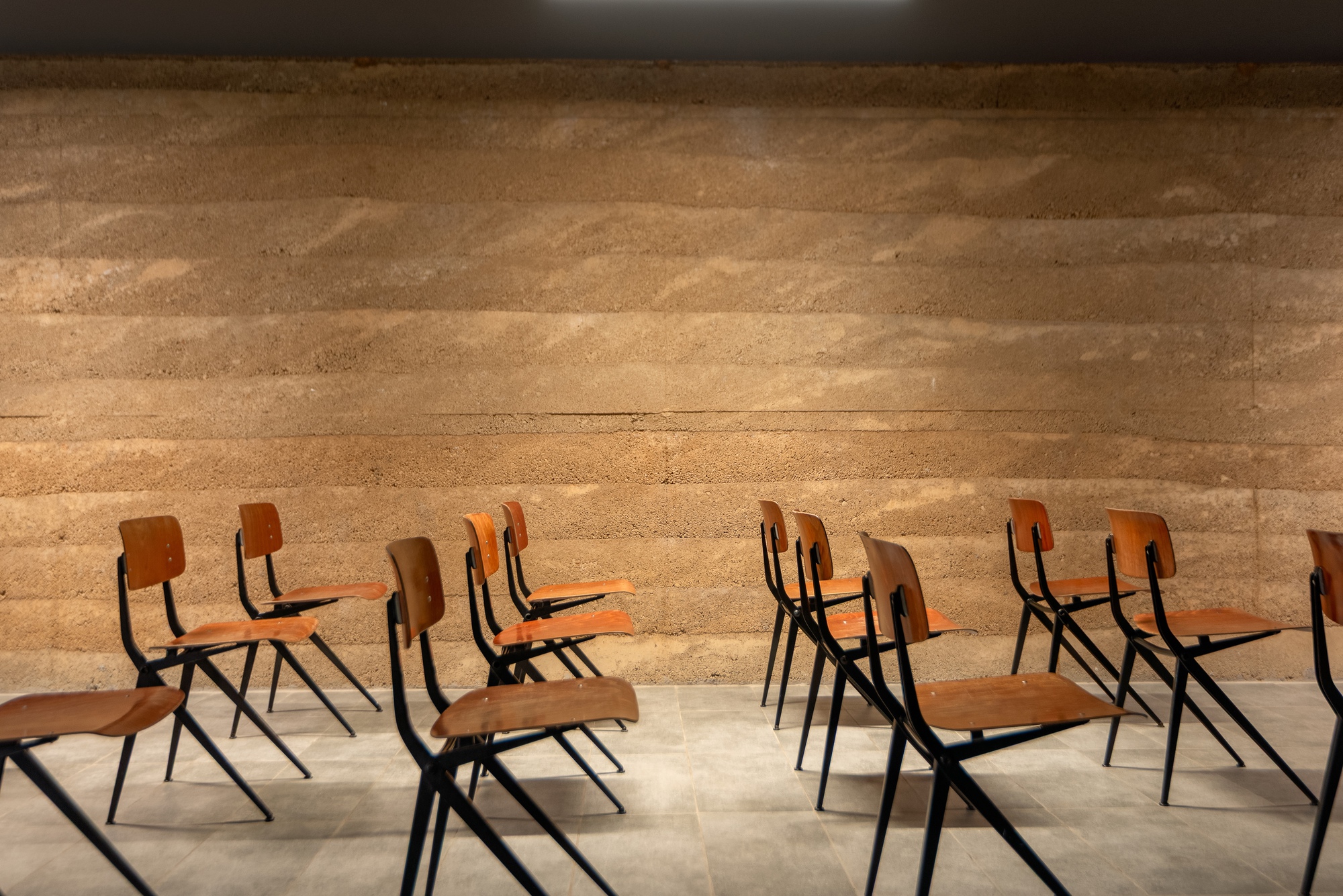 Row of wooden school chairs arranged before a horizontal layered rammed earth wall