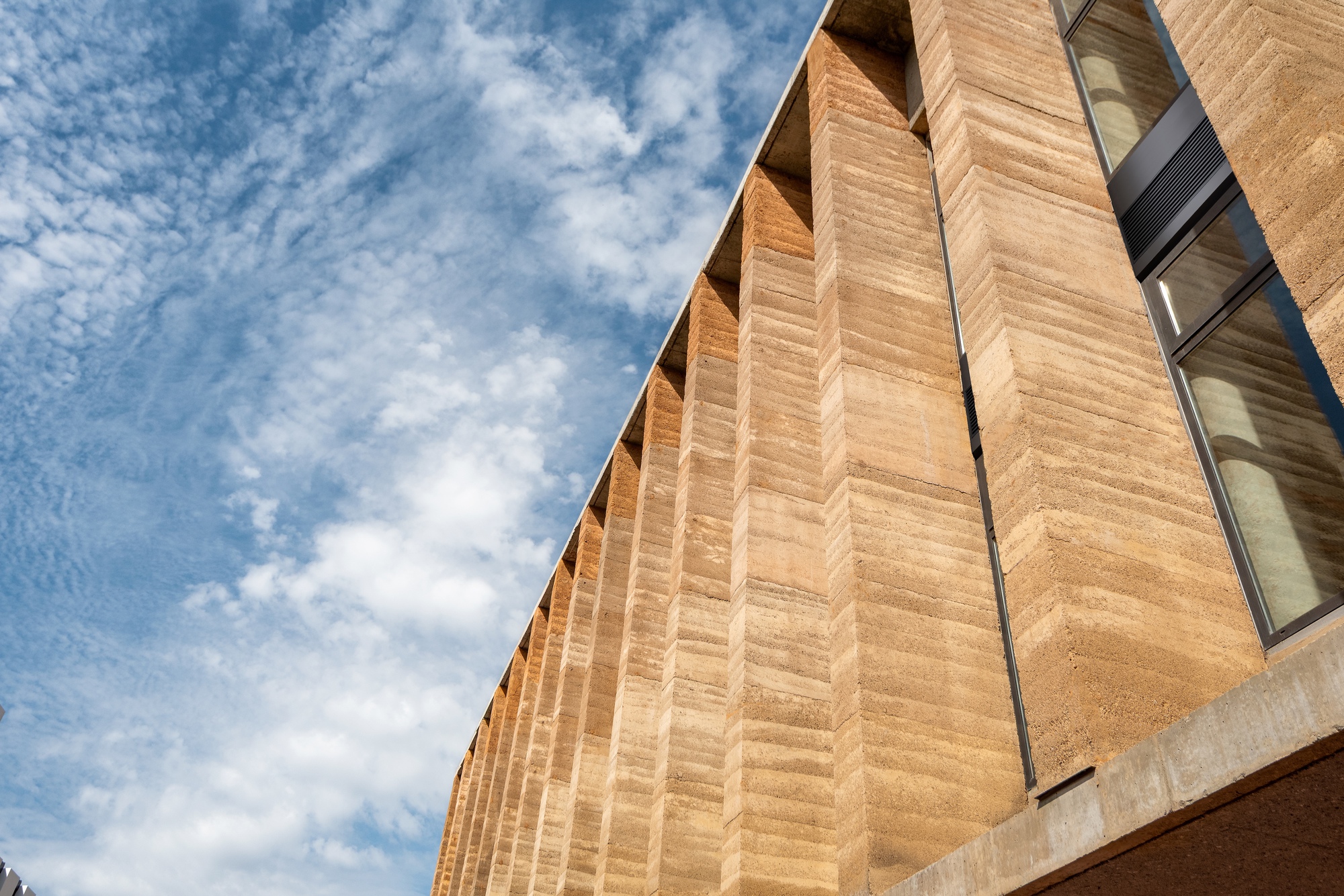Ribbed rammed earth facade with vertical fins casting shadows under a partly cloudy sky