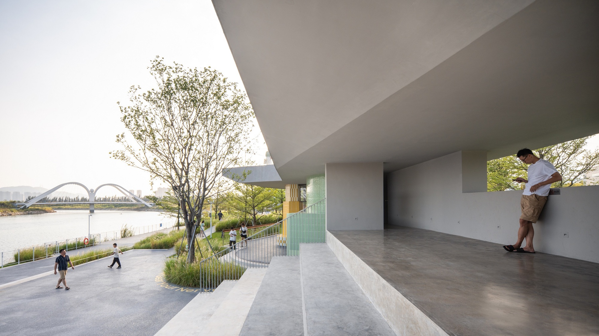 Covered terrace with concrete soffit overlooking a riverside promenade with pedestrians and trees
