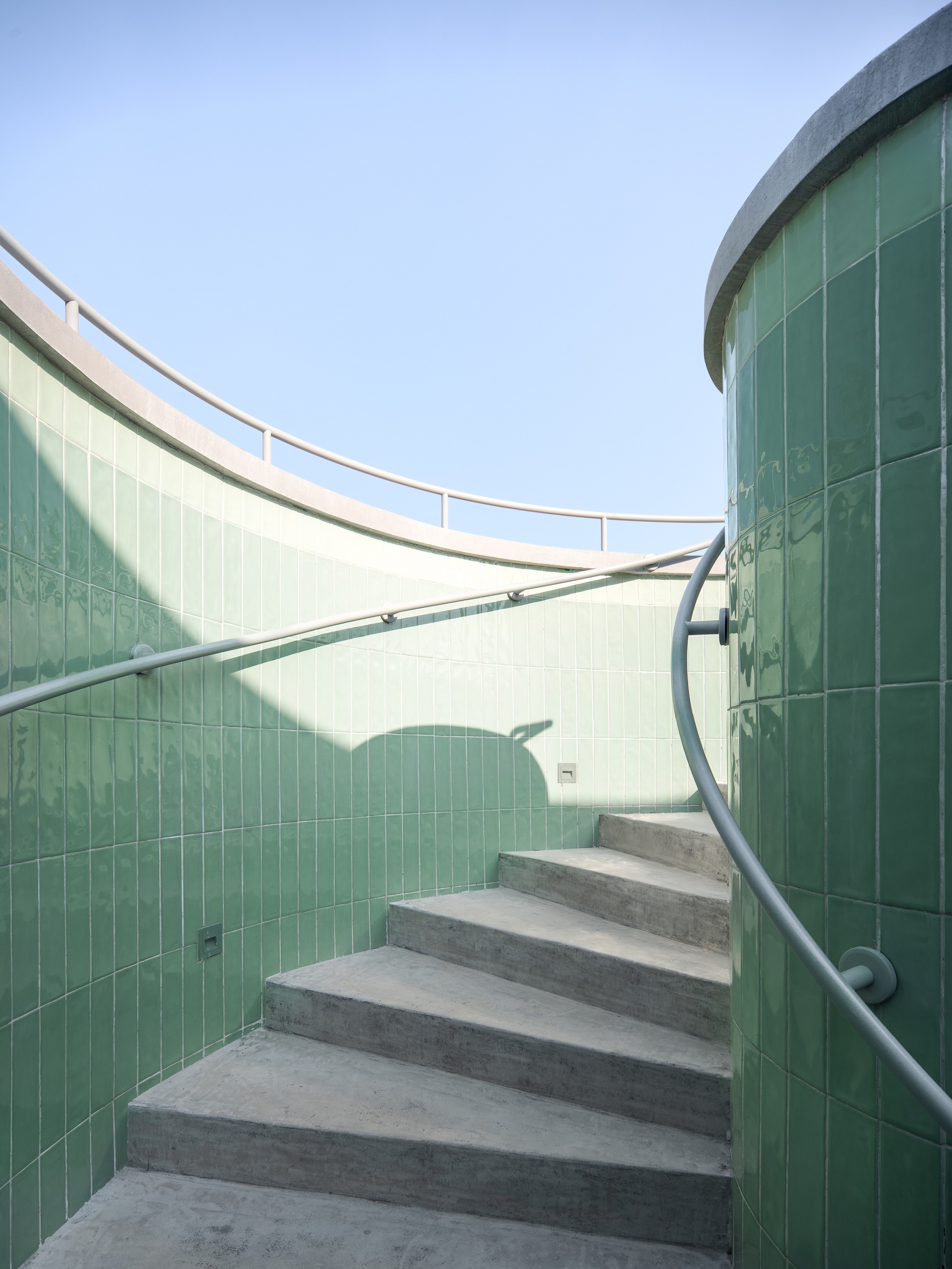 Curved concrete staircase with metal handrail ascending between green glass block walls under open sky