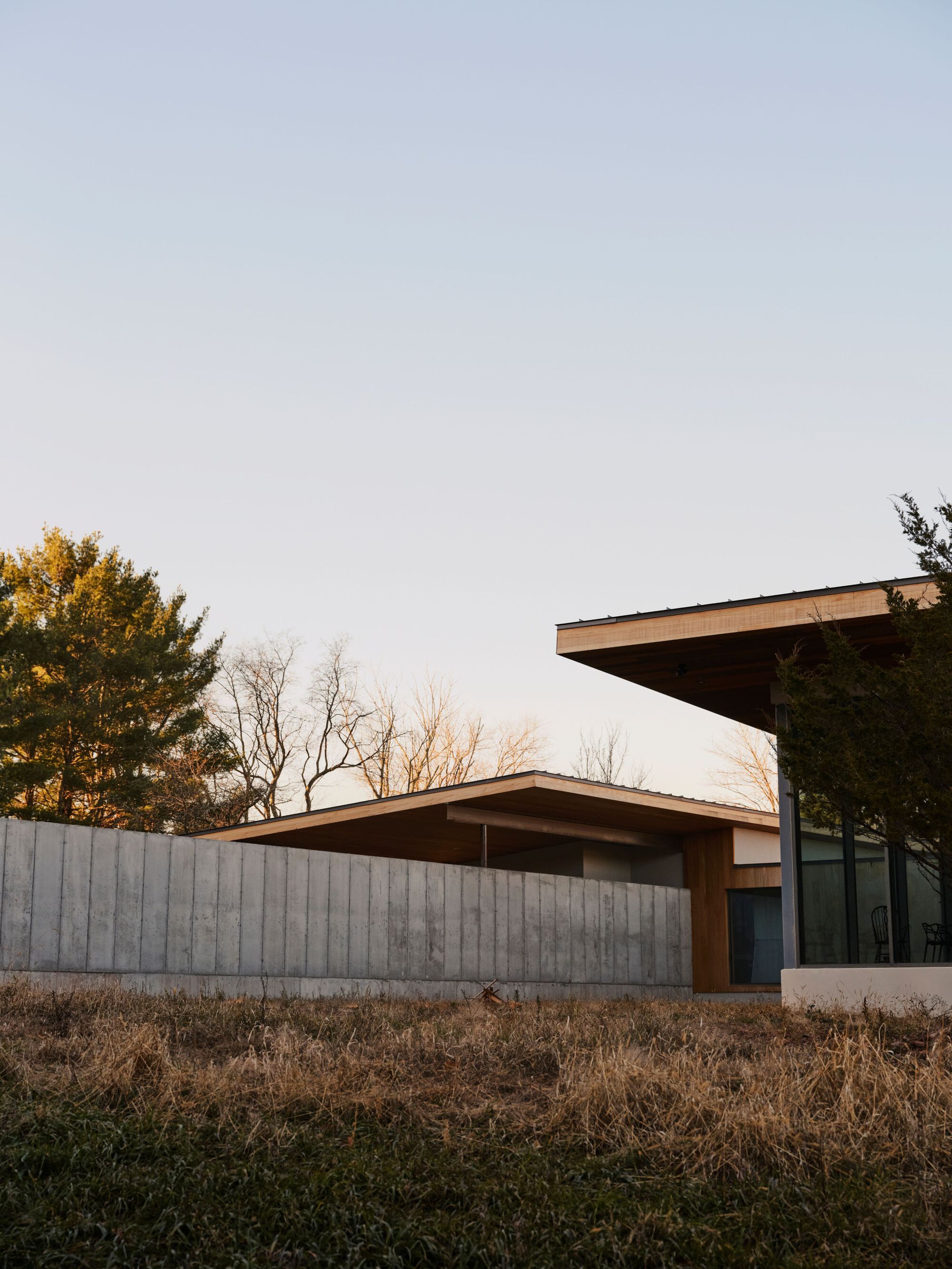 Cantilevered roof overhang above the board-formed concrete wall at dusk with bare trees beyond