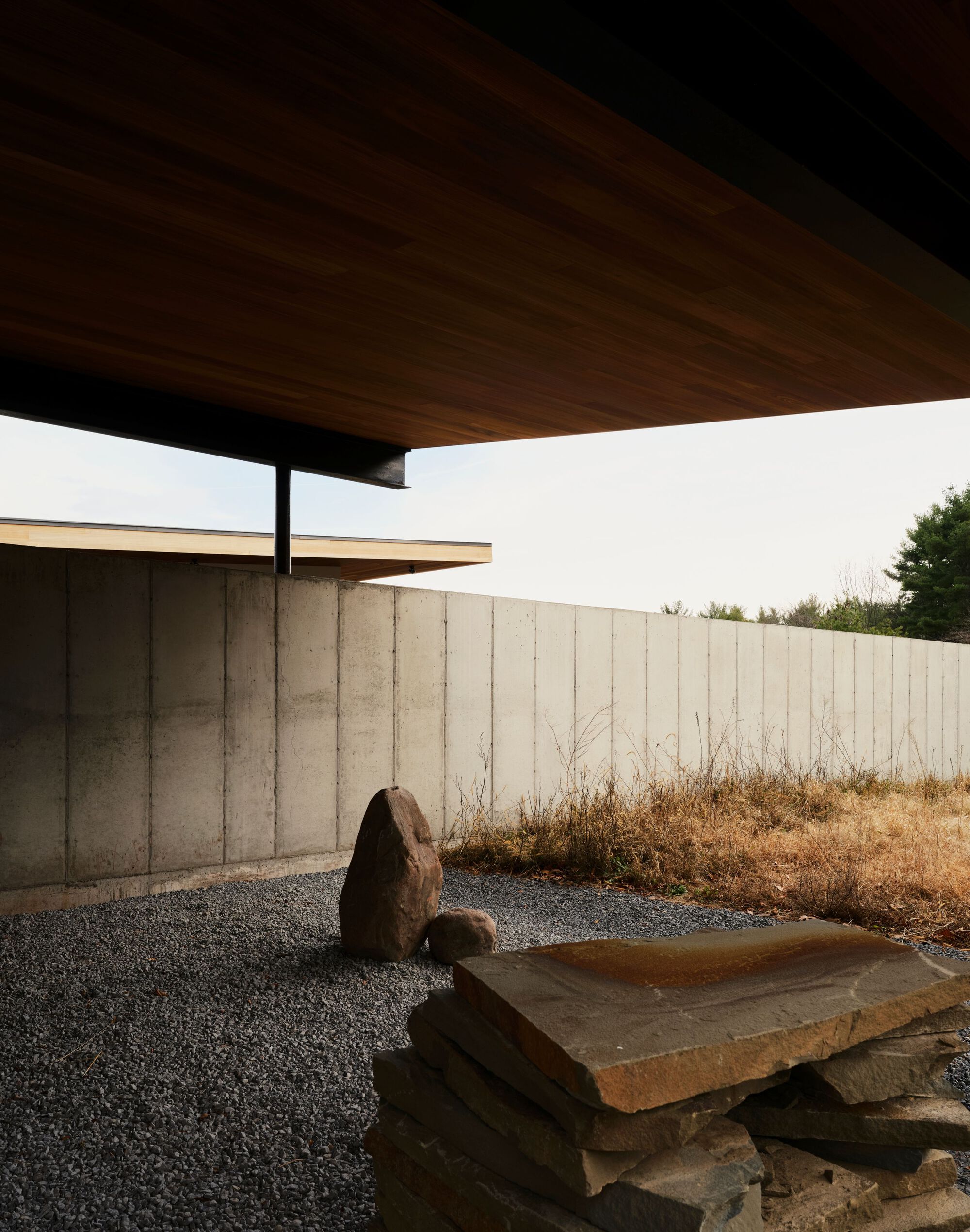Covered entry with timber soffit, concrete wall, gravel ground, and stacked stone water feature