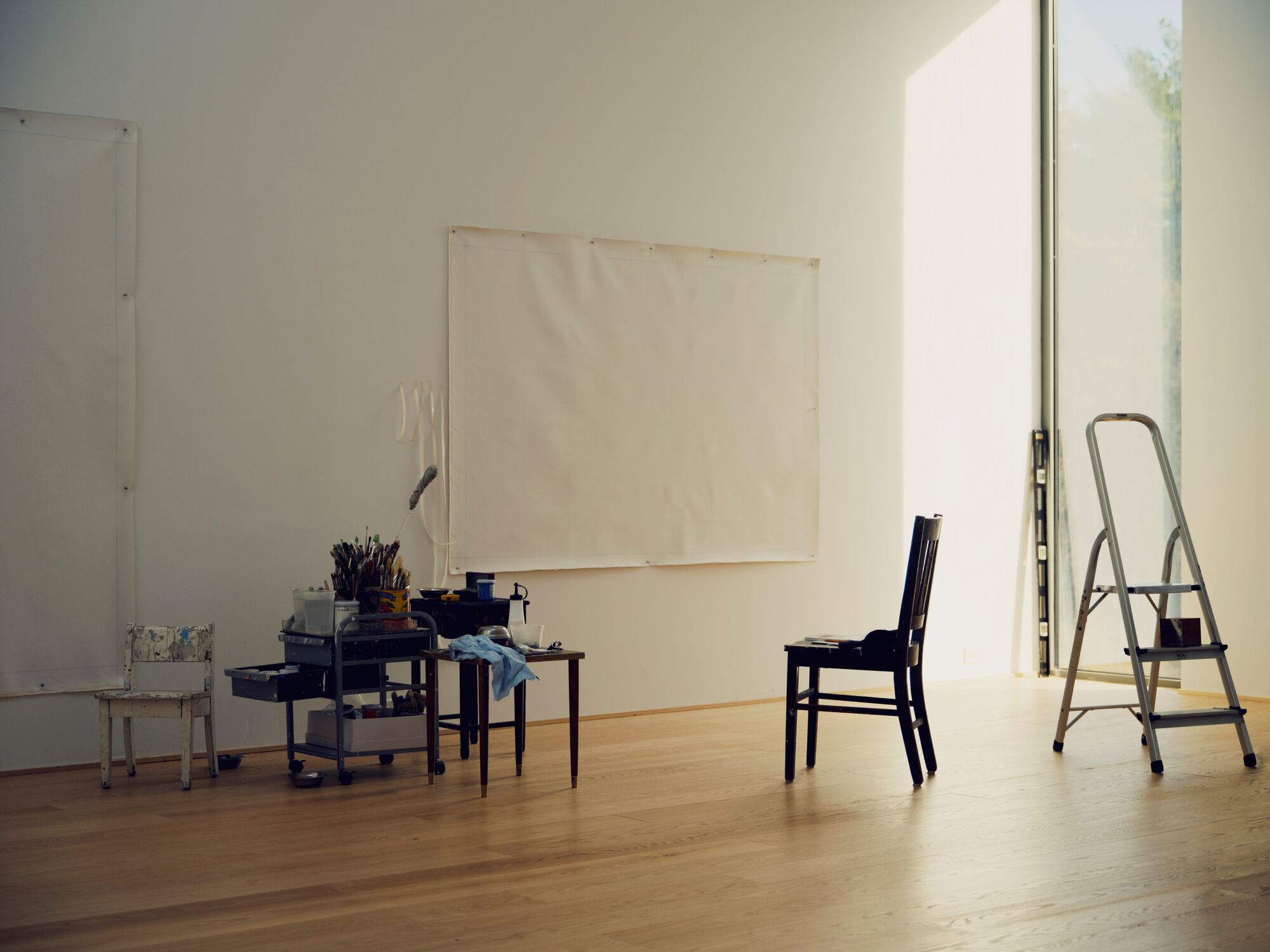 Corner of a workspace with blank canvas, supply cart and stepladder near a tall window