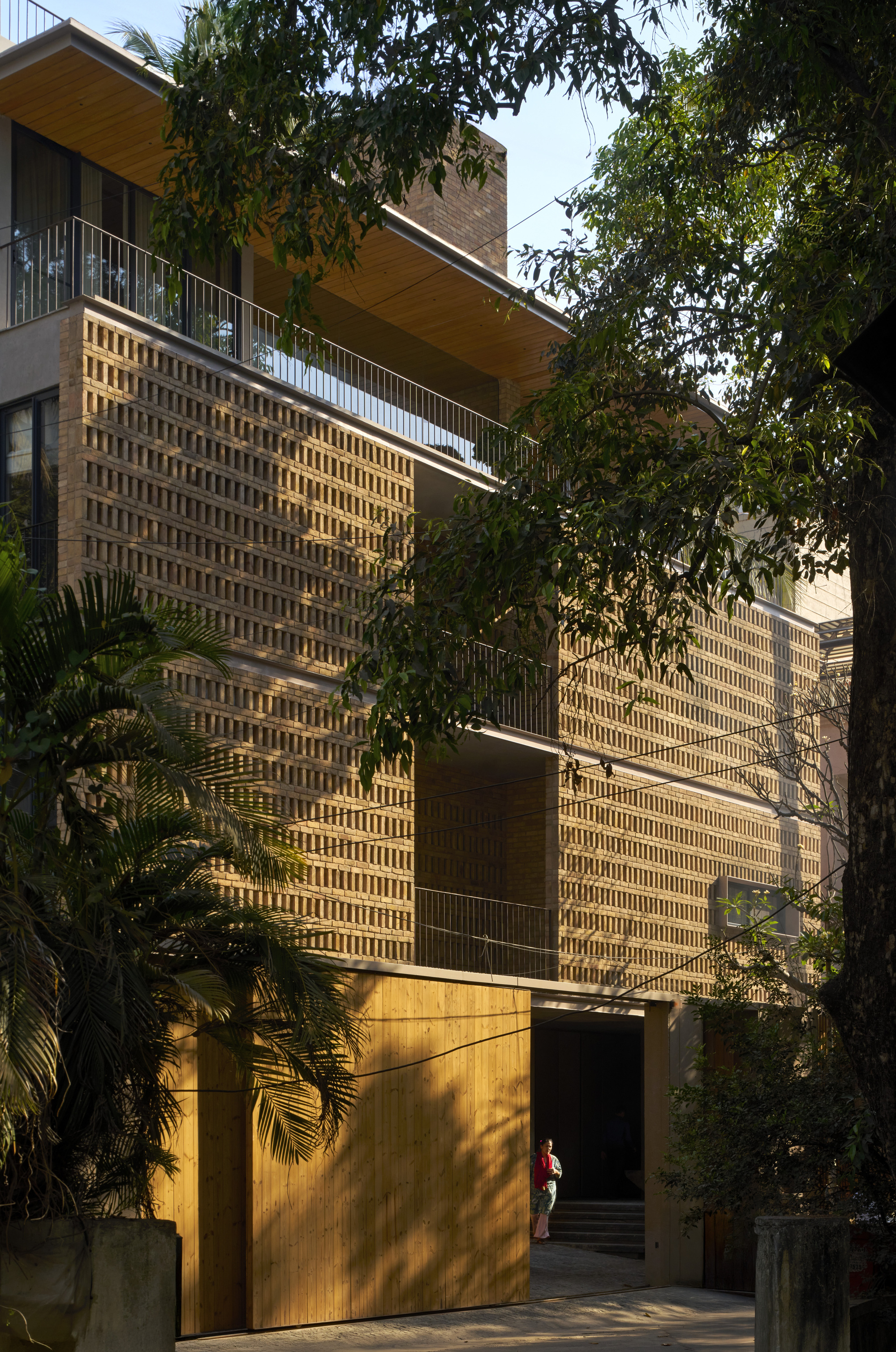 Street view of perforated brick screen facade behind palm fronds and dappled afternoon light