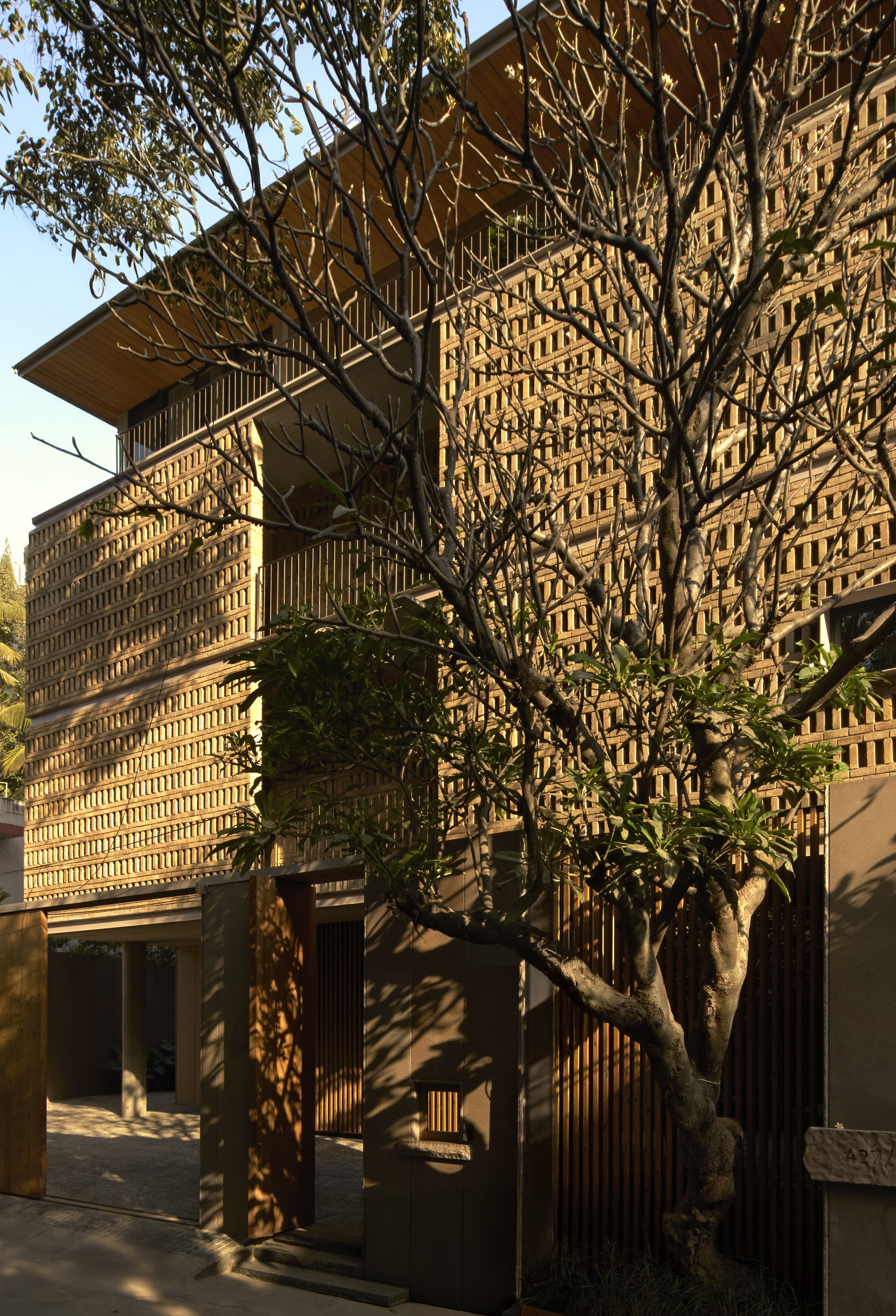 Layered perforated brick facade with deep overhangs behind a bare winter tree in afternoon light
