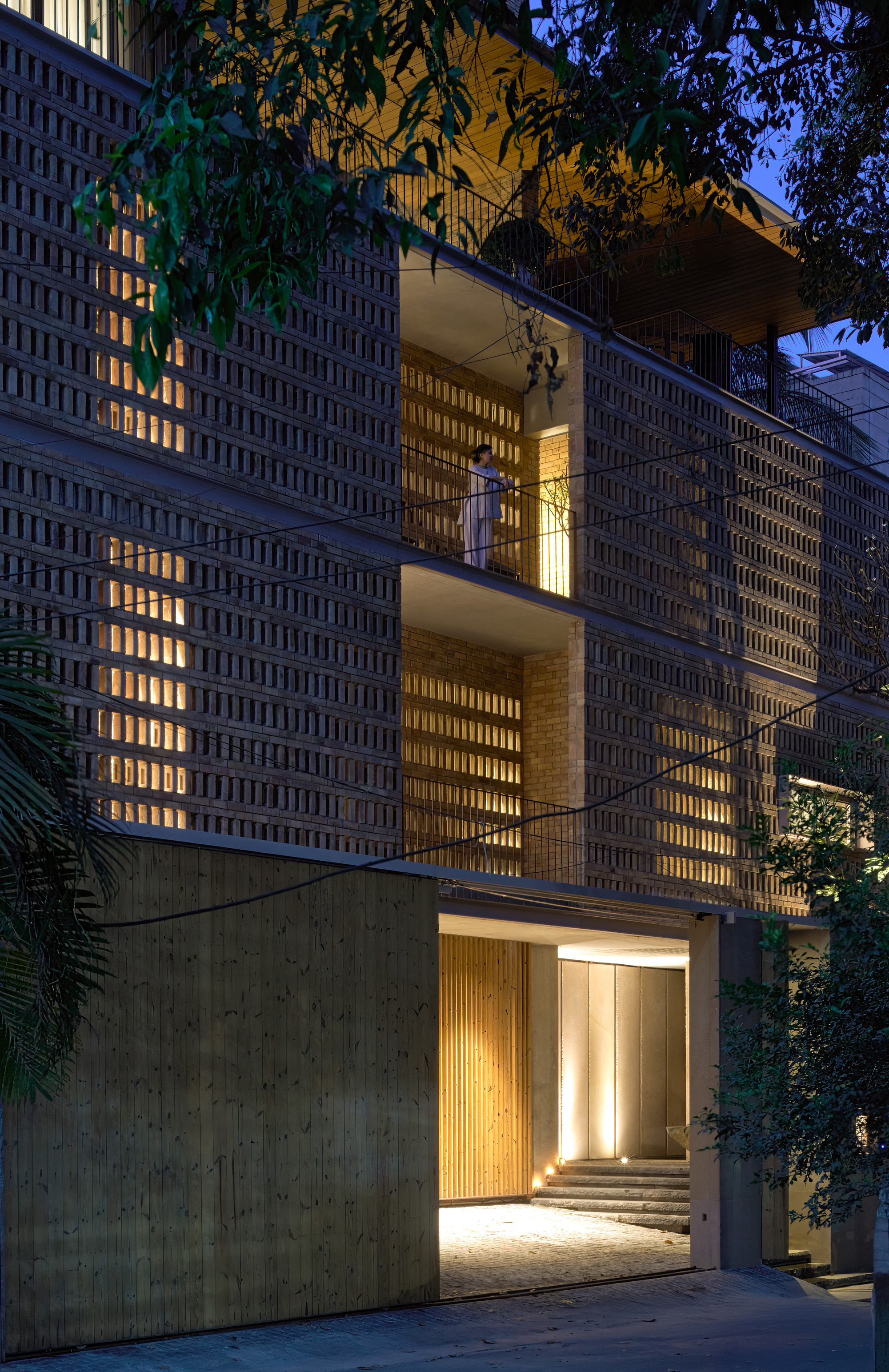 Illuminated perforated brick facade with stacked balconies and entrance at dusk surrounded by vegetation