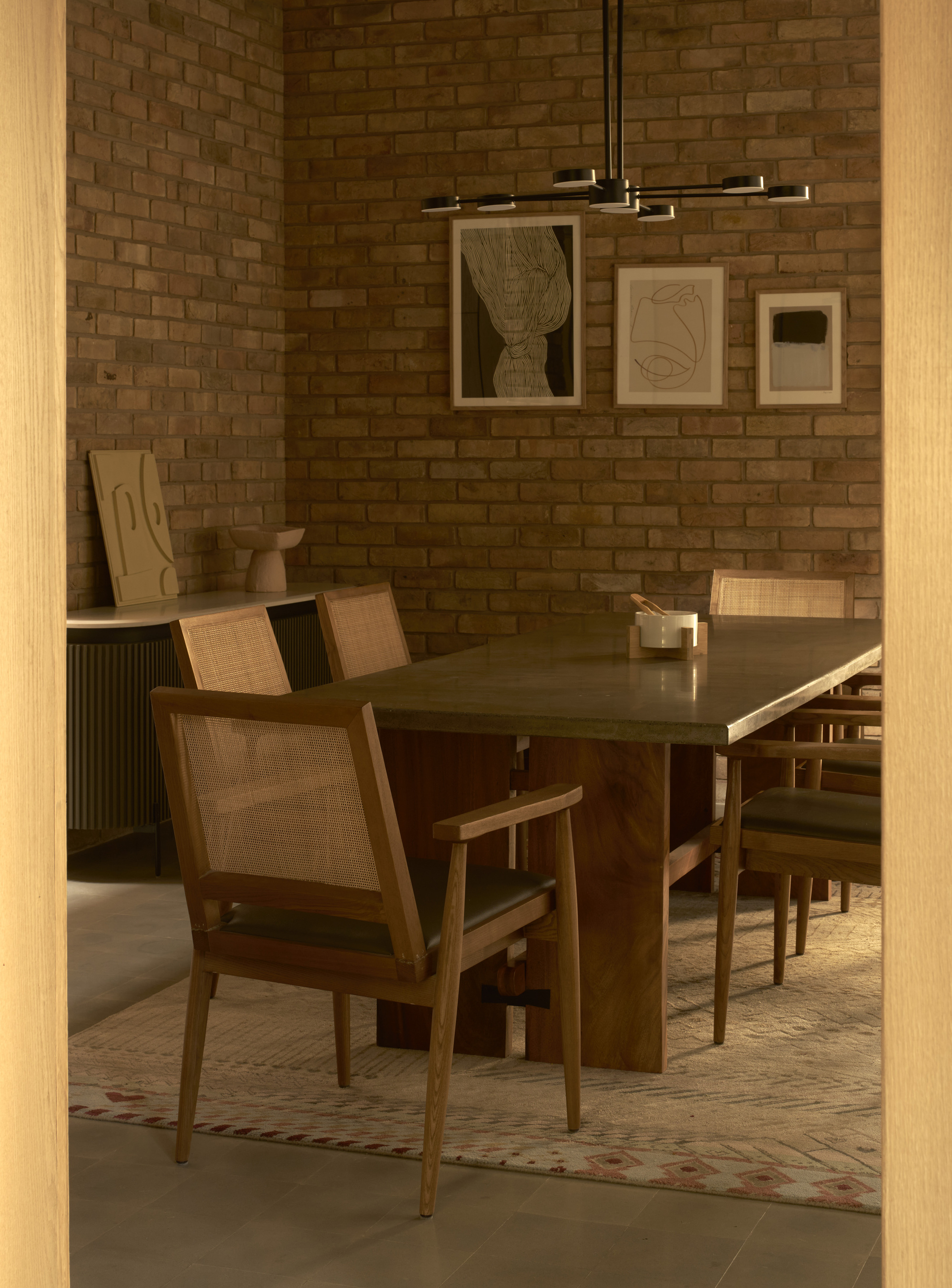 Dining room with brick wall, timber table and chairs, and framed artworks lit by pendant fixture