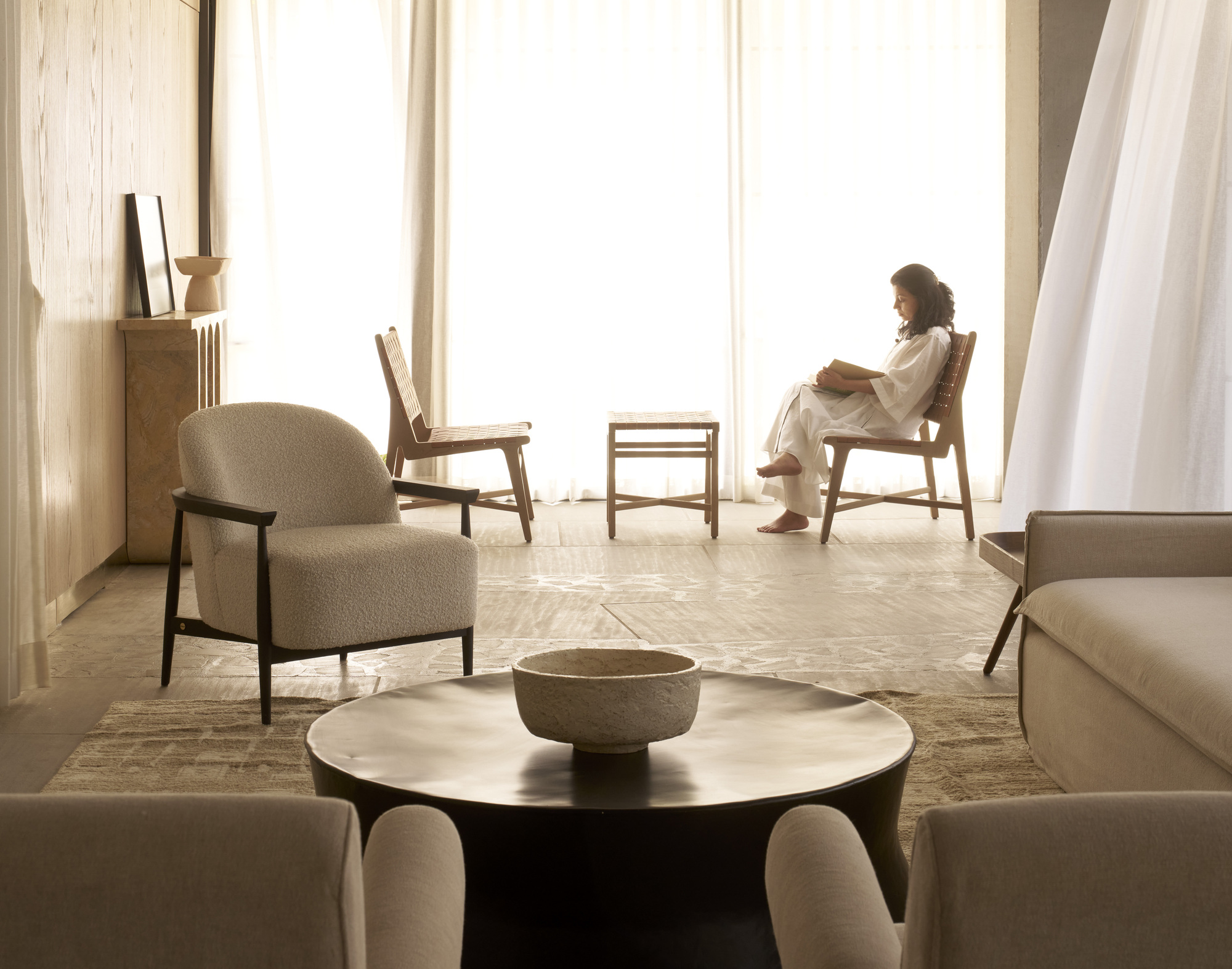 Living space with seated figure by floor-to-ceiling windows in soft daylight