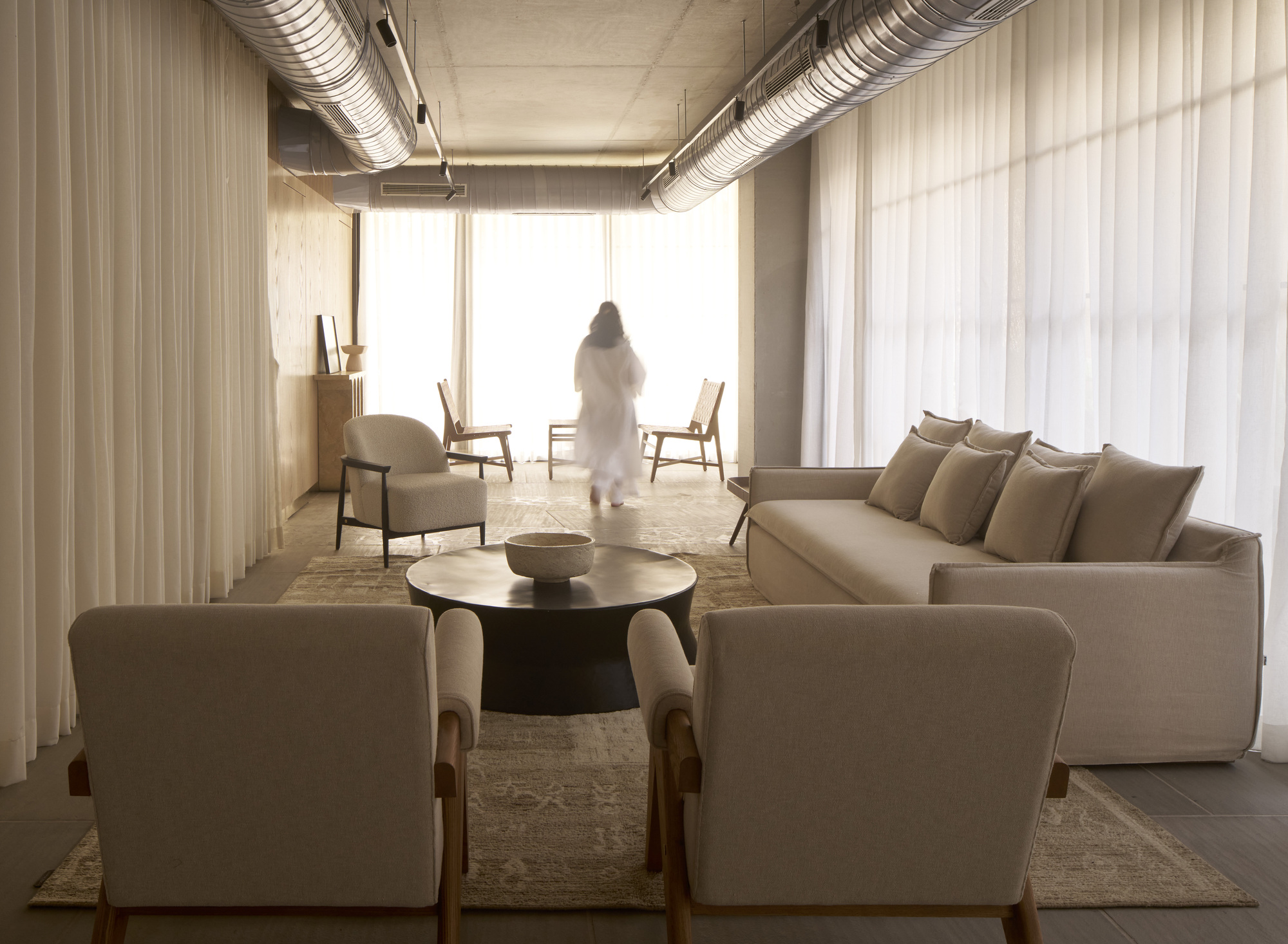 Living area with exposed ceiling ducts, sheer curtains and a figure silhouetted against bright windows