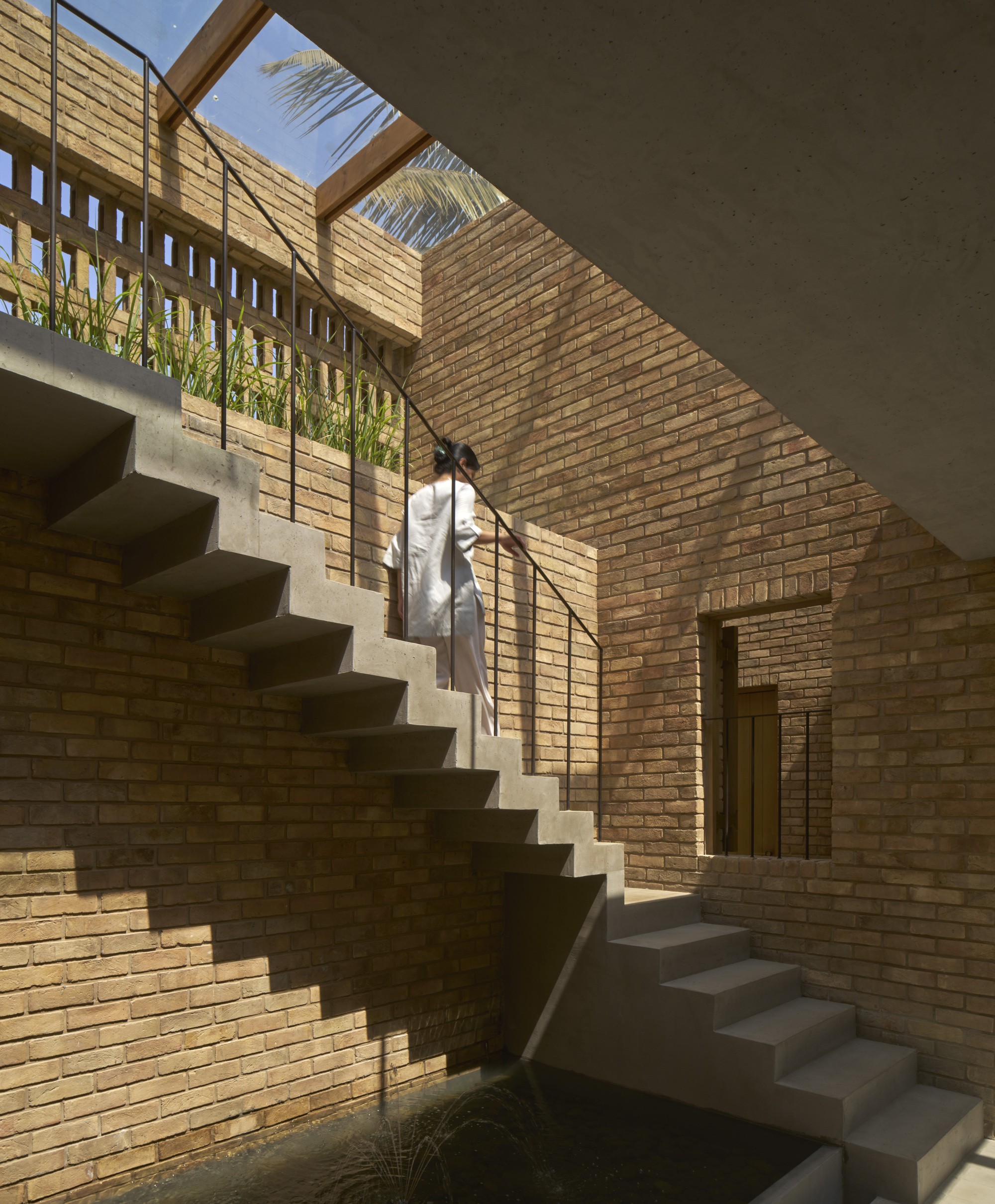 Concrete staircase rising through a brick courtyard with clerestory openings and a figure ascending