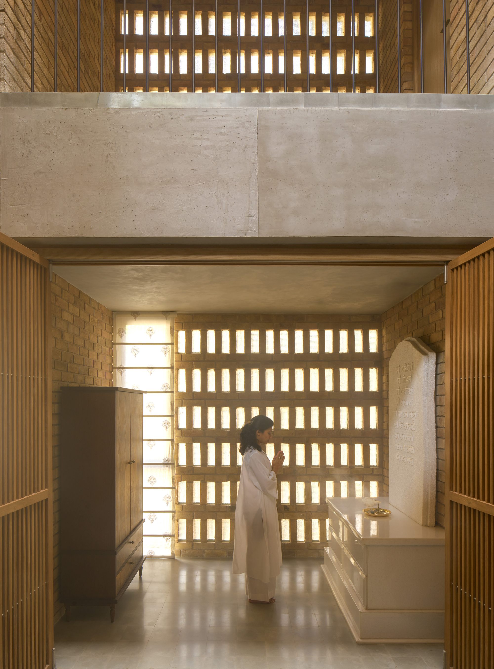 Double-height kitchen with gridded brick screen wall and figure standing at counter beneath concrete mezzanine