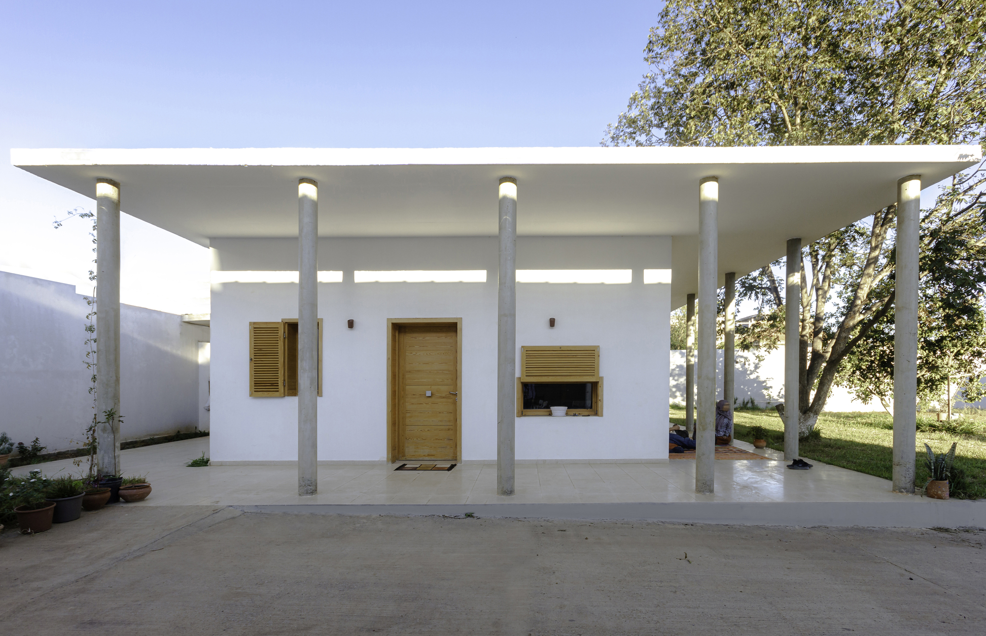 White stucco volume with wood shutters and central door beneath a flat canopy supported by concrete columns