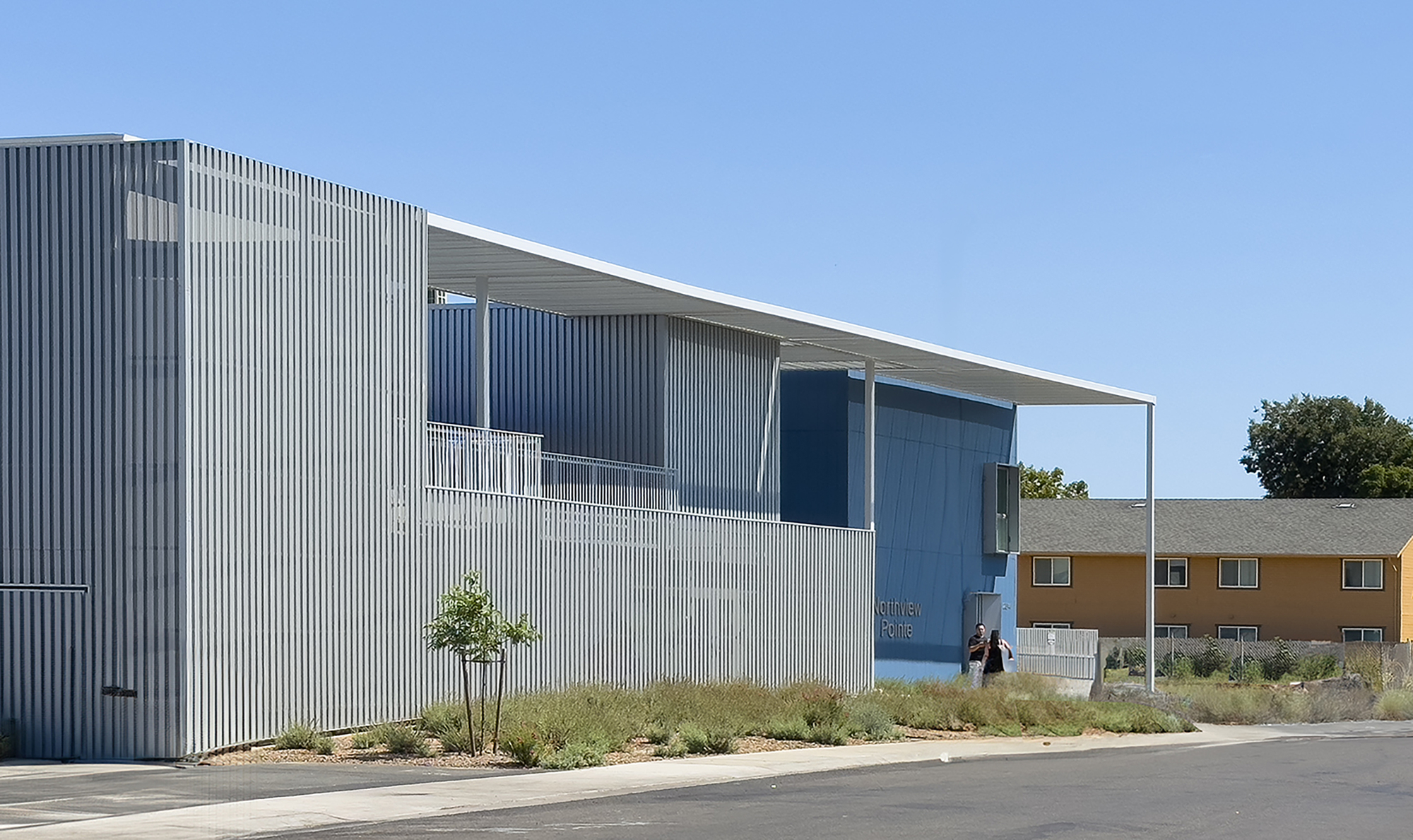 Oblique view of the corrugated metal facade with planted grasses along the sidewalk in sunlight