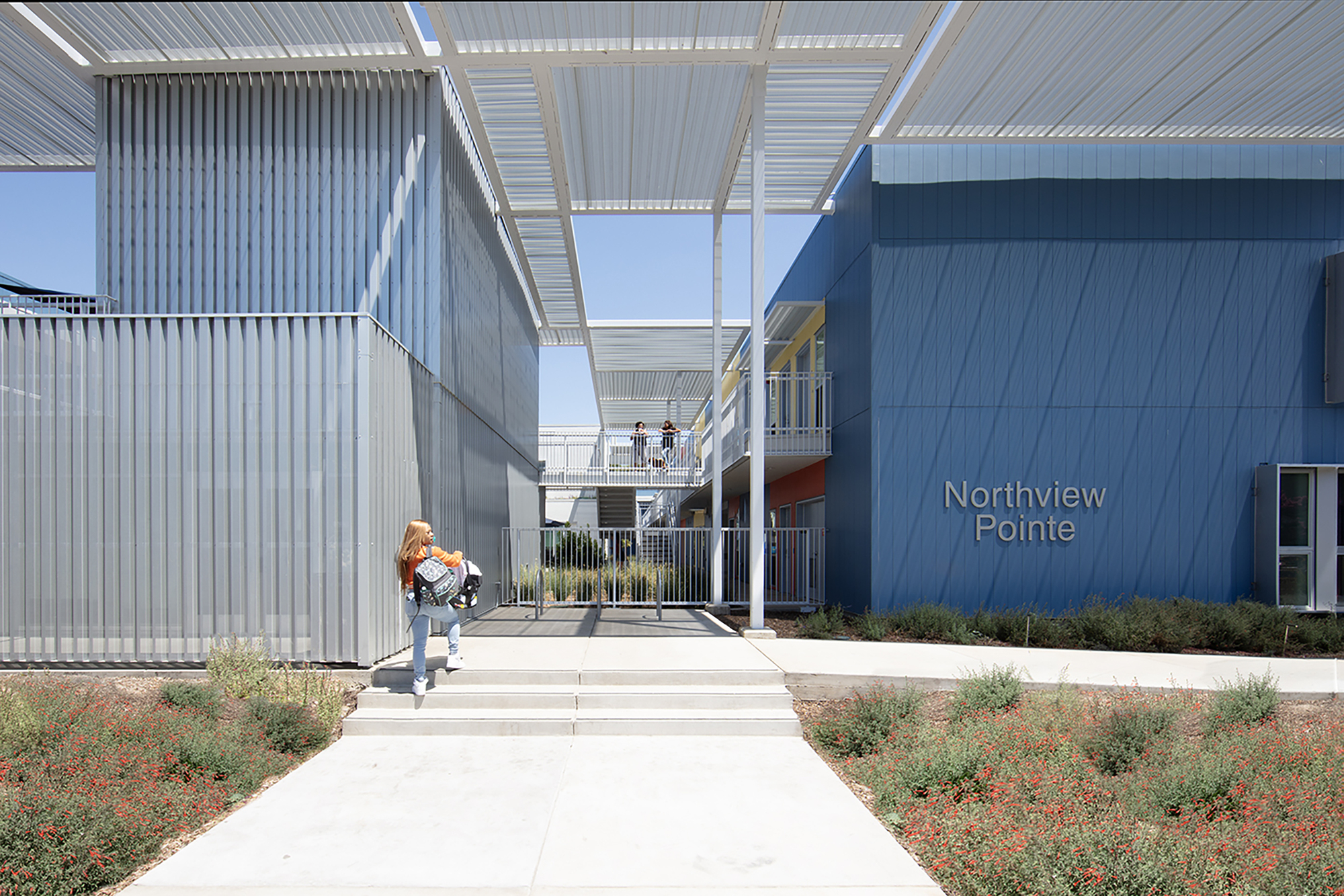 Central courtyard walkway with corrugated metal volumes and white canopy overhead under clear skies