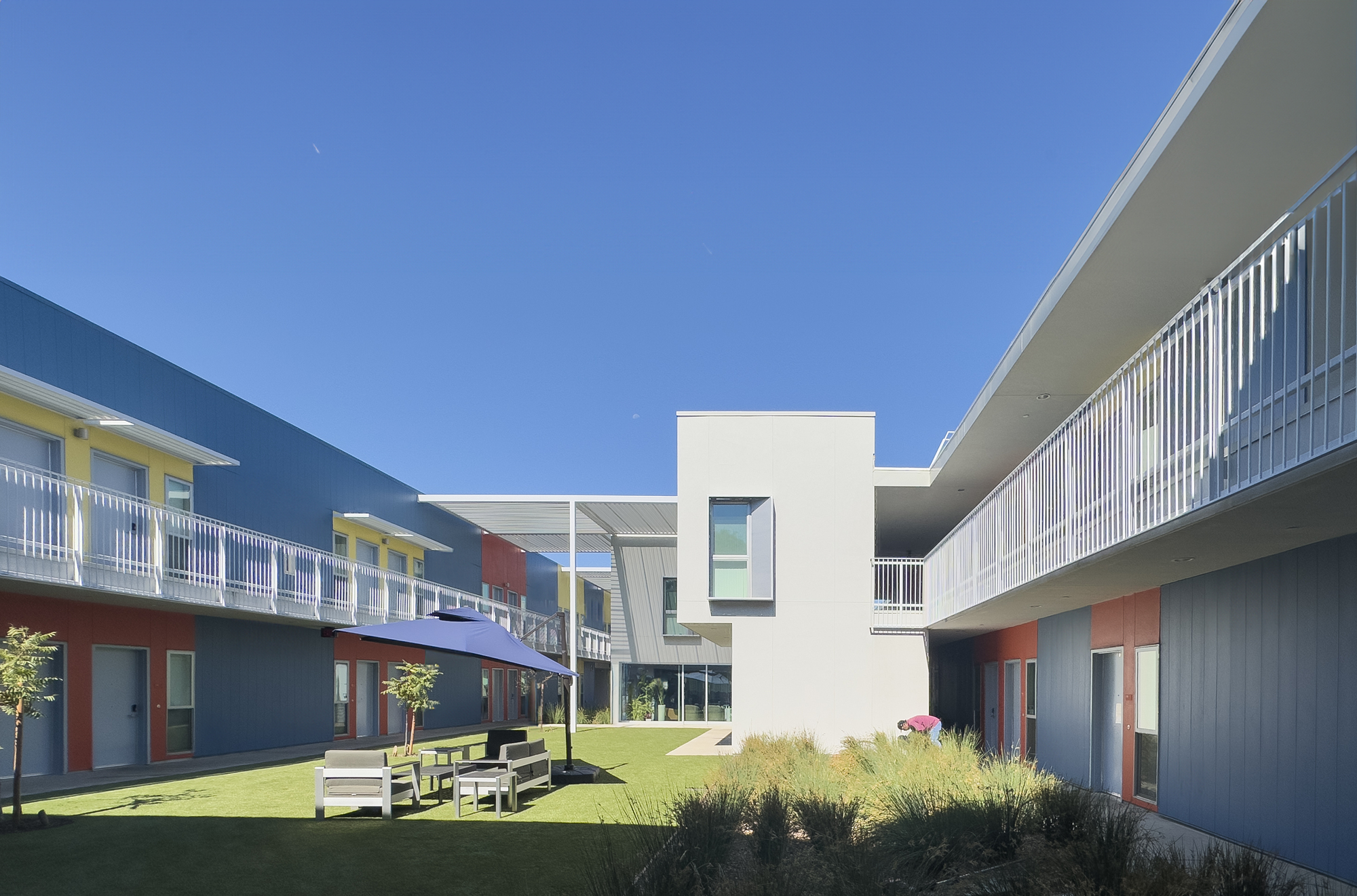 Courtyard view framed by white corrugated volumes and double-loaded corridors with outdoor benches on grass
