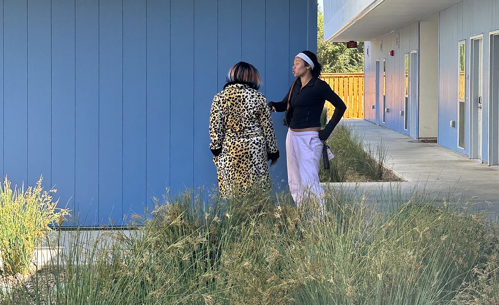 Two people conversing beside the blue panel facade and landscape buffer at the covered walkway edge