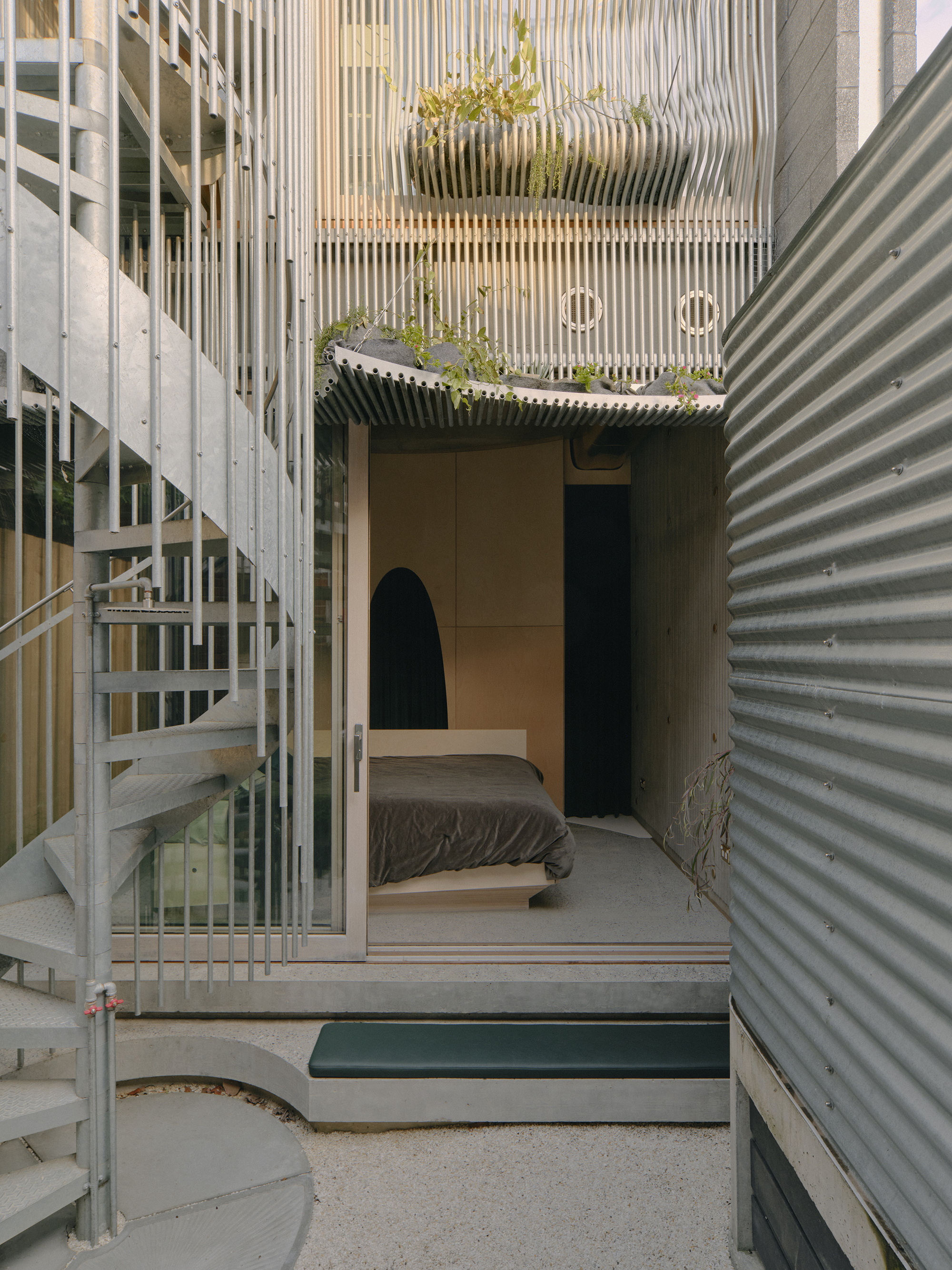 Courtyard view through vertical timber screen showing planted bed above corrugated metal walls and spiral stair