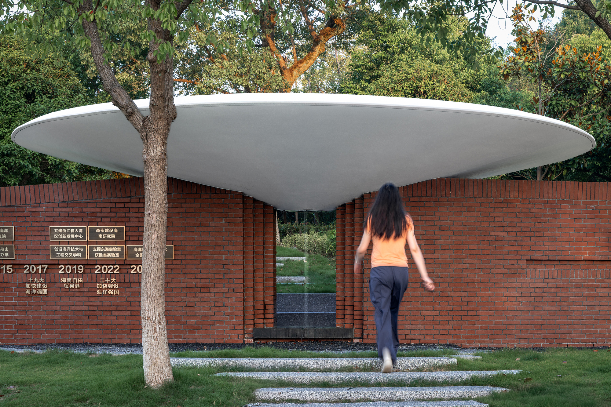 Red brick wall memorial with white disc roof canopy and visitor approaching along grass path