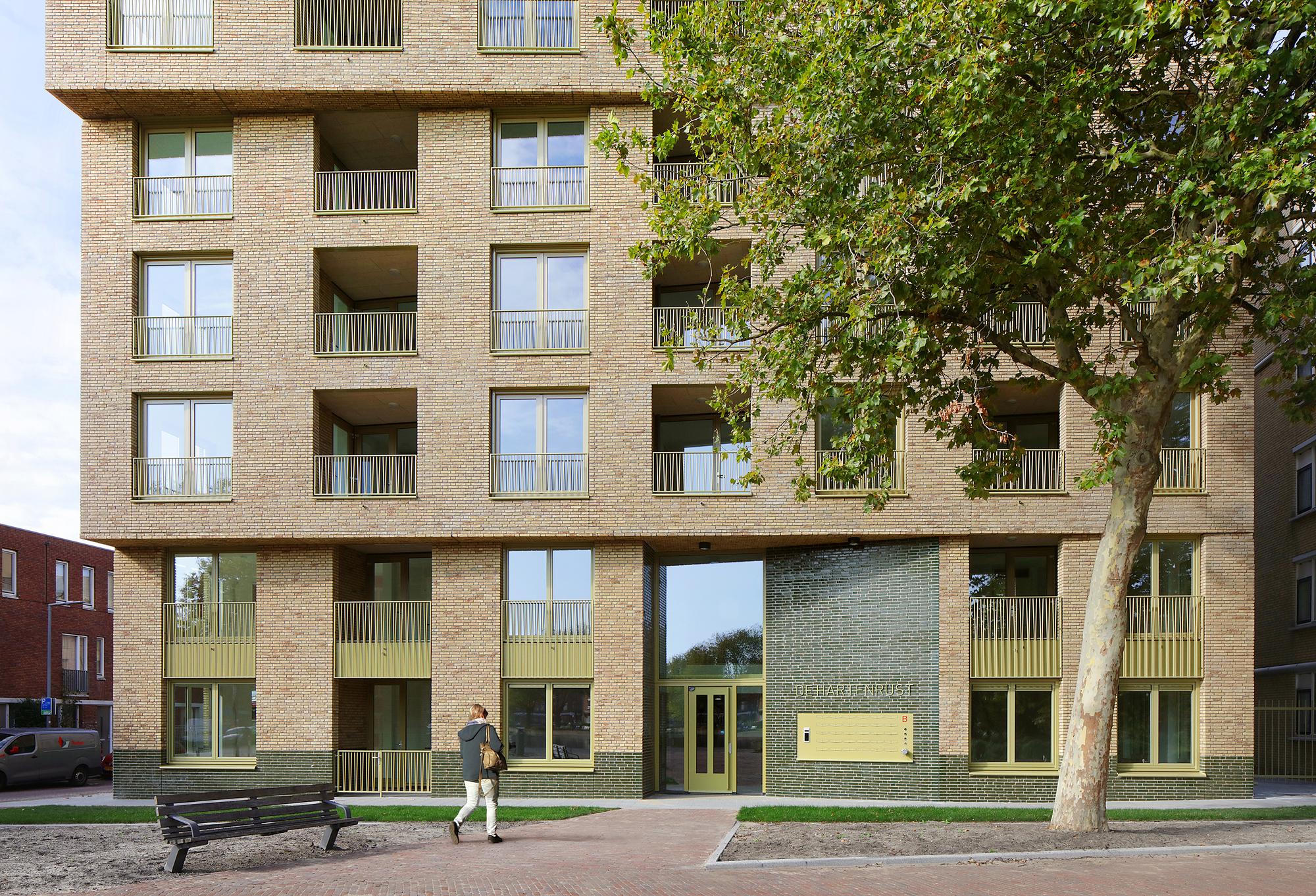 Ground-level facade showing recessed balconies with metal railings and a pedestrian walking past a bench
