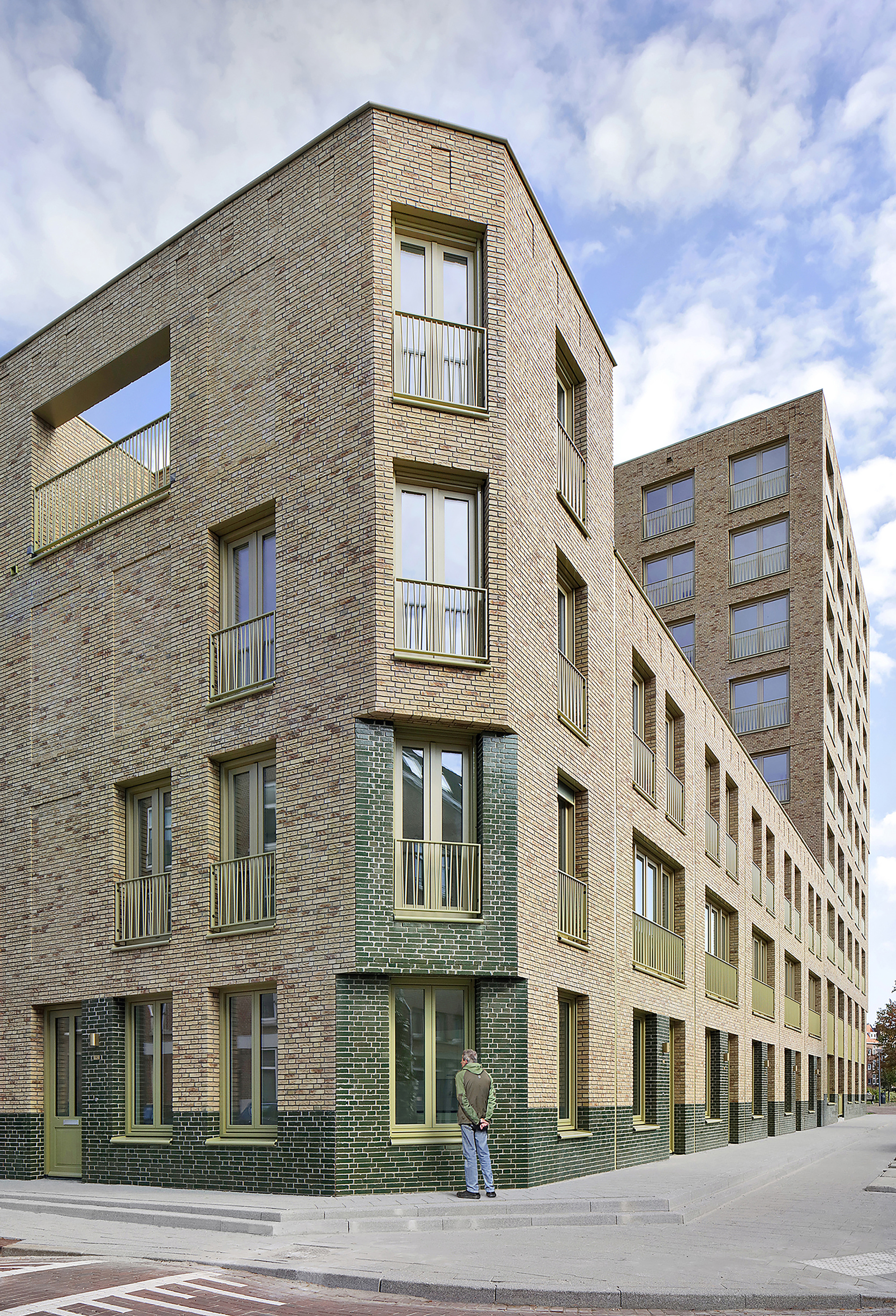 Chamfered corner of a buff brick residential building with metal balconies and a pedestrian below